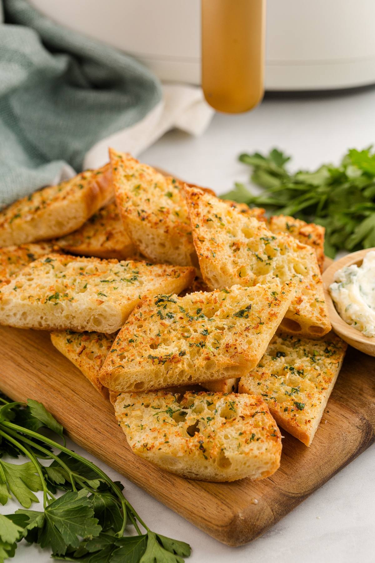 Sliced garlic bread with herbs on a wooden board, surrounded by fresh parsley and a bowl of dip.