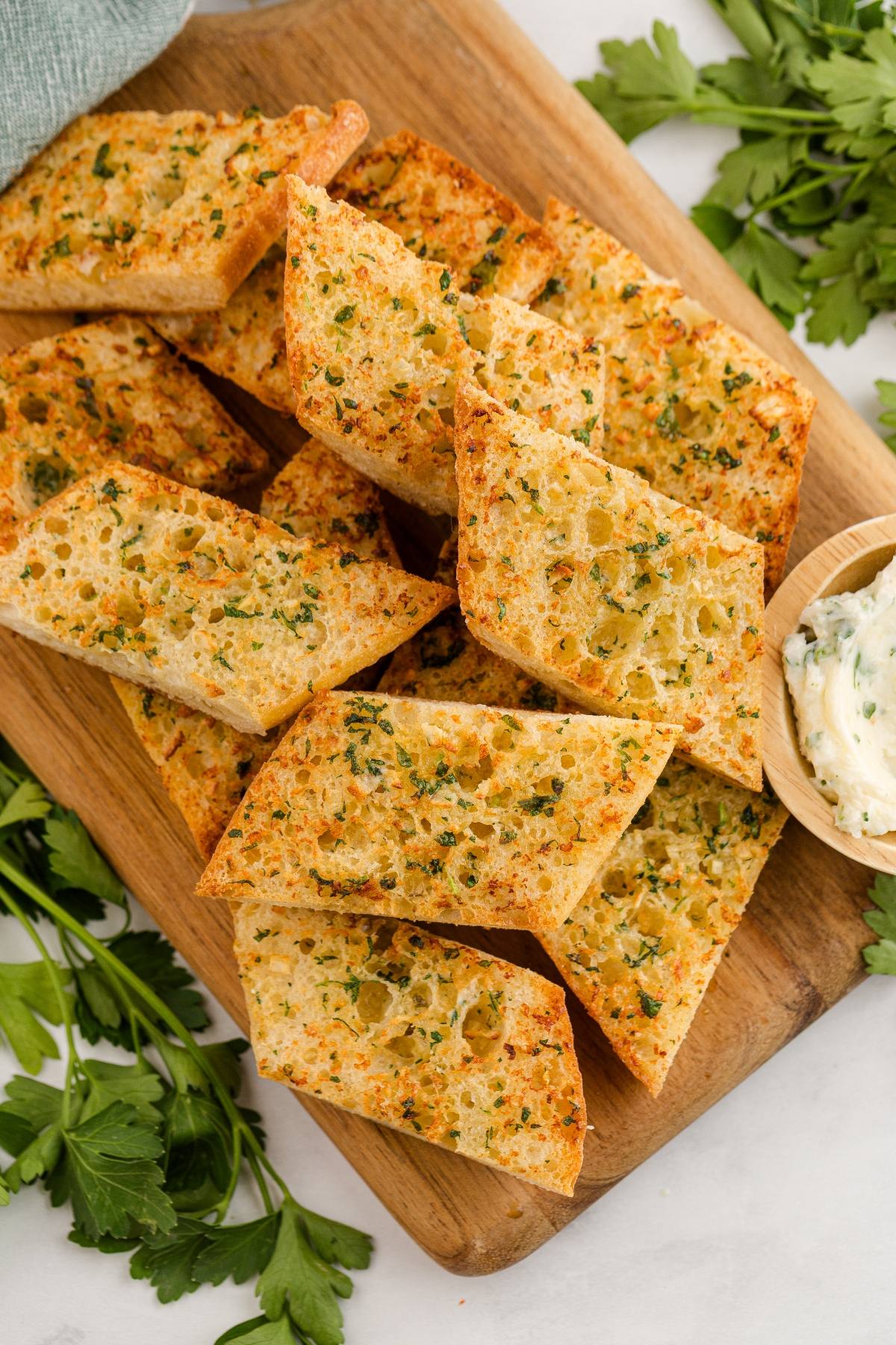 Slices of golden, crispy garlic bread topped with herbs on a wooden board, with fresh parsley nearby.