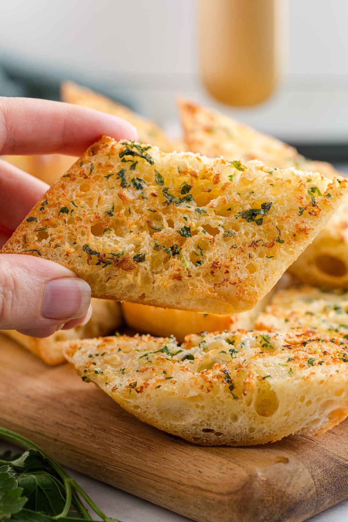 A hand holds a piece of crispy garlic bread topped with herbs, with more slices on a wooden board.