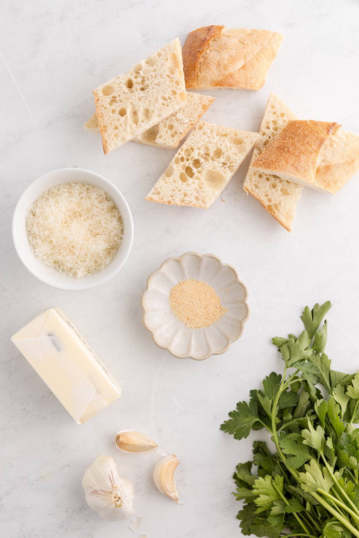 Sliced bread, grated cheese, garlic, parsley, butter, and garlic powder arranged on a white surface.