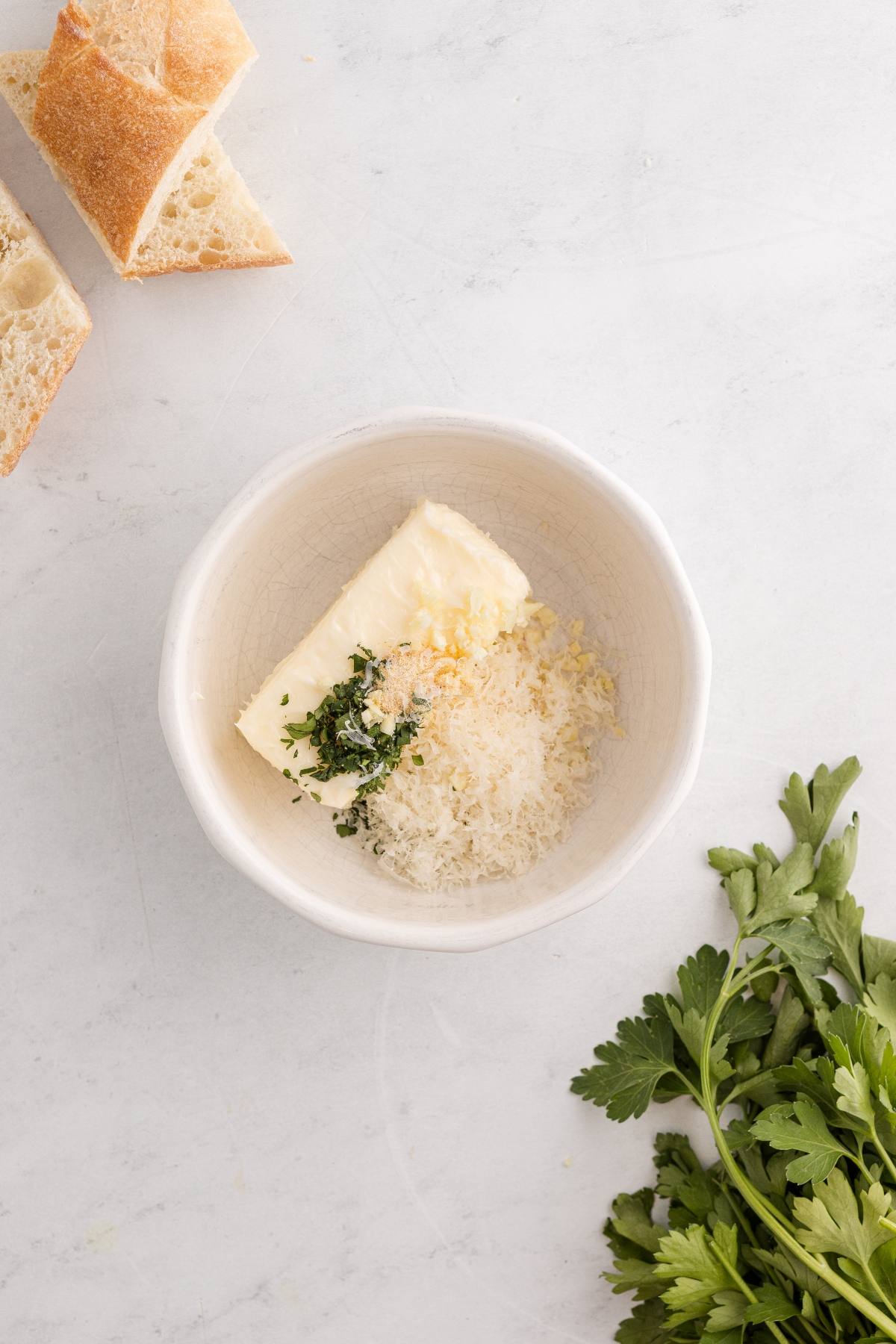 A bowl with butter, grated cheese, chopped herbs, and garlic, with bread slices and parsley nearby.