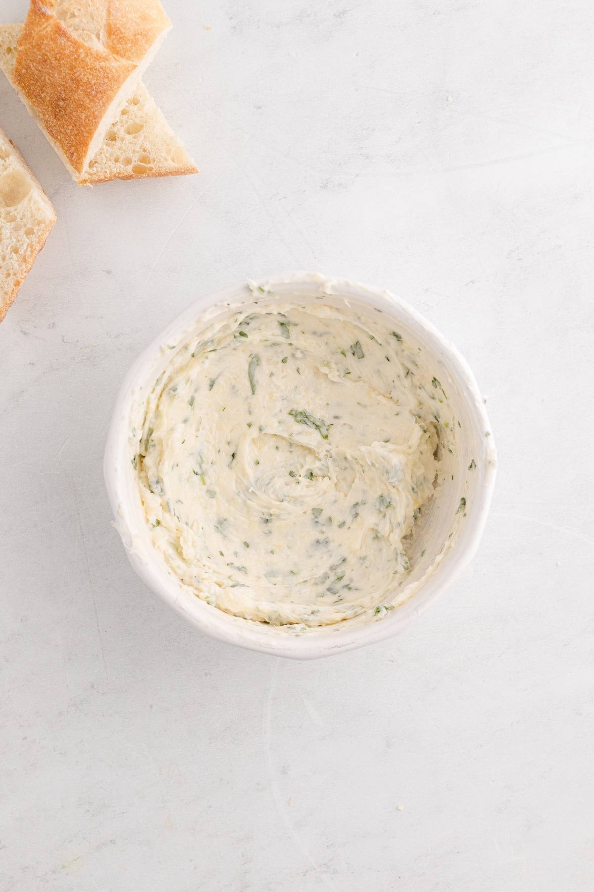 White bowl of herb butter next to sliced pieces of bread on a light-colored surface.