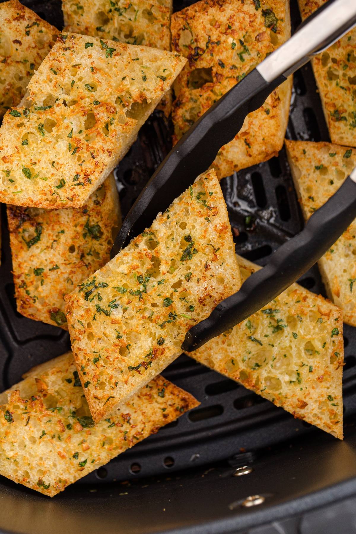 Tongs holding a piece of golden, crispy garlic bread over more slices in an air fryer basket.