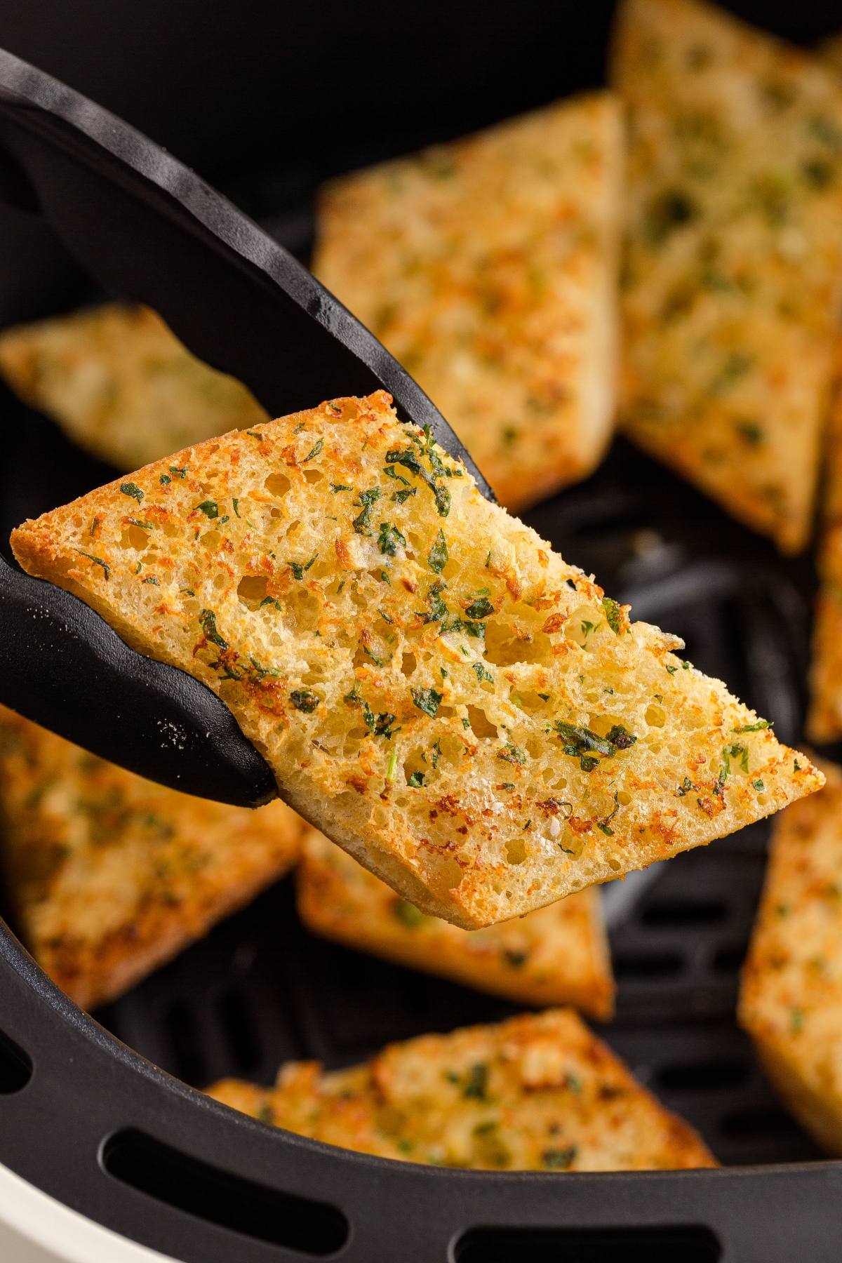 A close-up of garlic bread being held with tongs over more garlic bread slices in an air fryer basket.