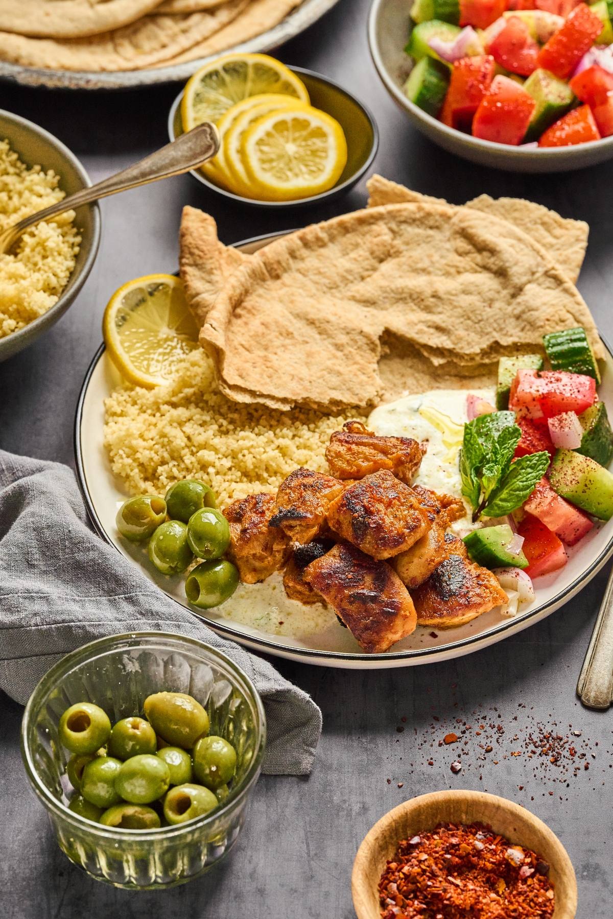 Plate with grilled chicken, couscous, pita bread, olives, salad, lemon slices, and yogurt sauce.