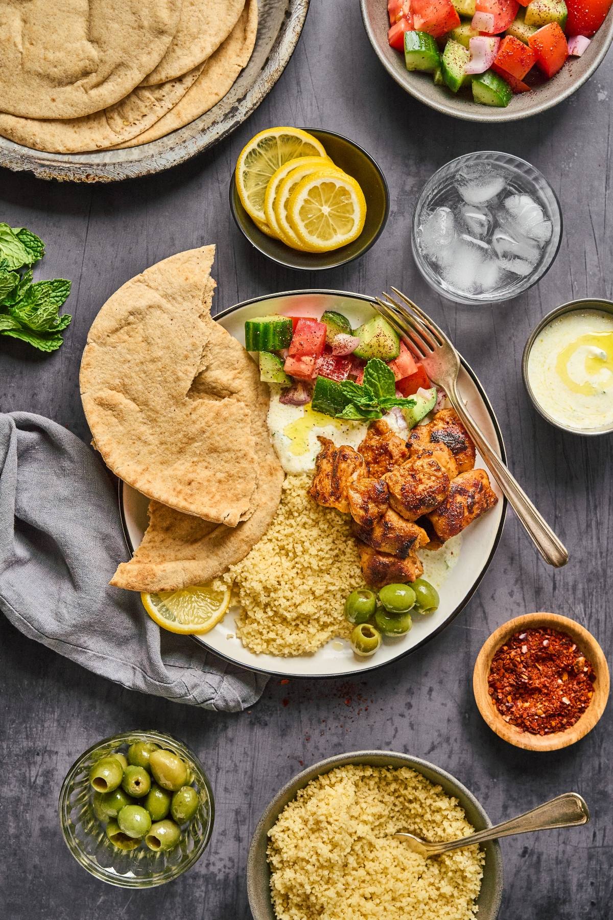 A plate with pita, grilled chicken, couscous, olives, salad, and lemon slices, surrounded by side dishes.