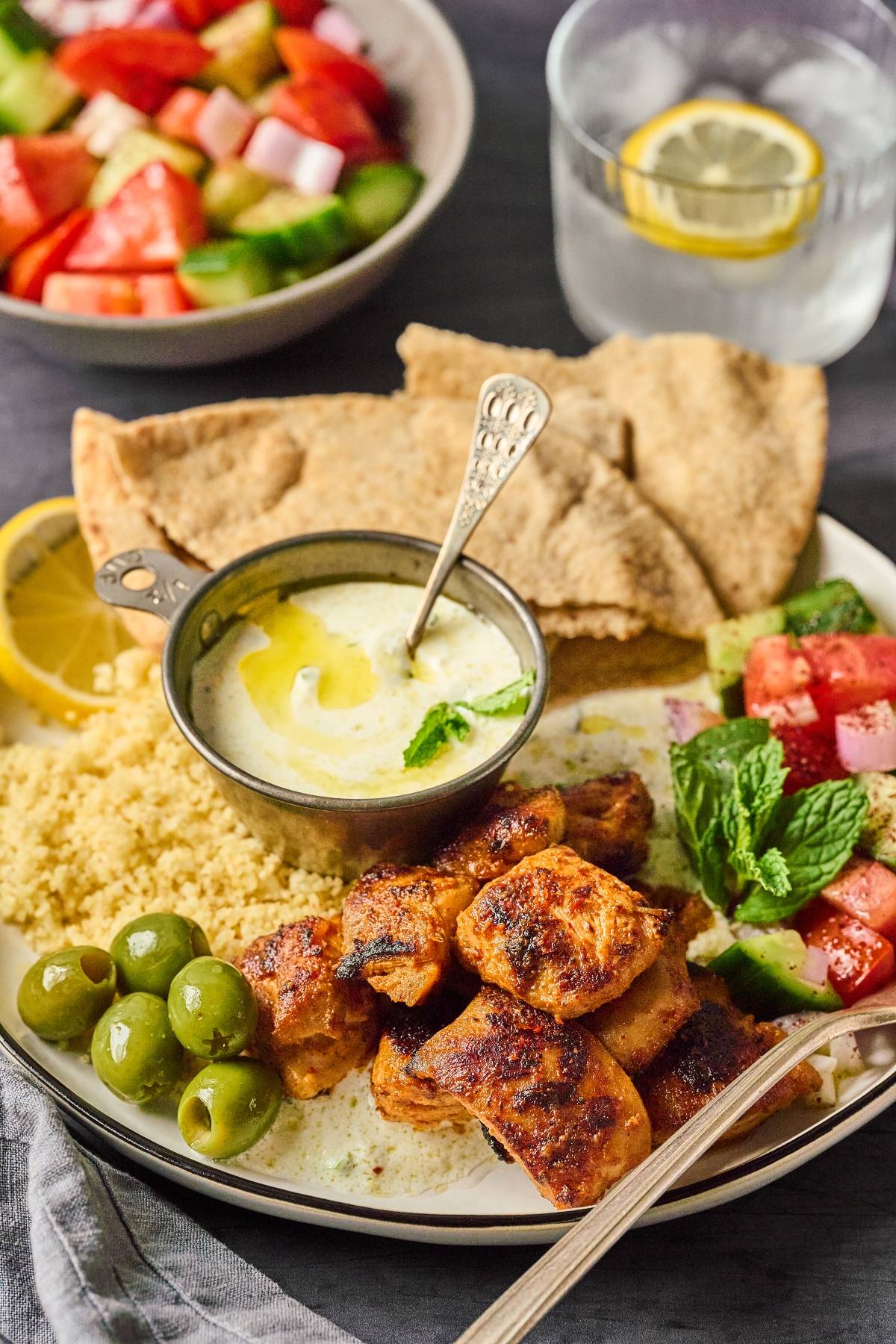 A plate with grilled chicken, couscous, olives, salad, pita bread, and a bowl of yogurt sauce.
