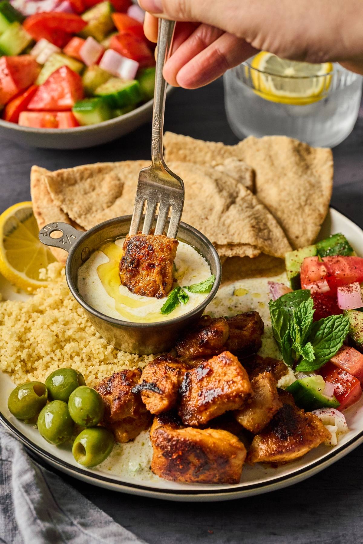 A hand dips spiced grilled chicken into sauce, with couscous, pita, olives, and salad on the plate.