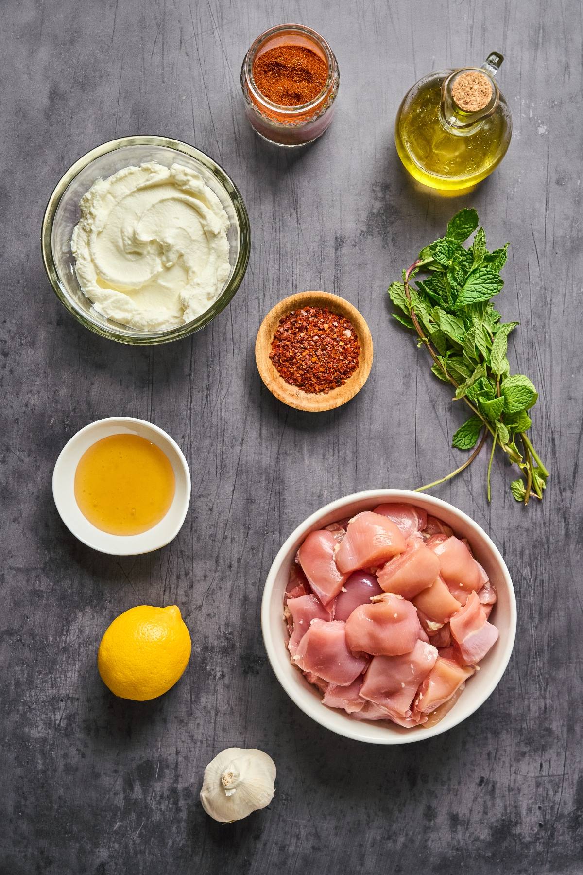 Bowls of yogurt, spices, oil, honey, herbs, lemon, garlic, and diced raw chicken on a gray surface.