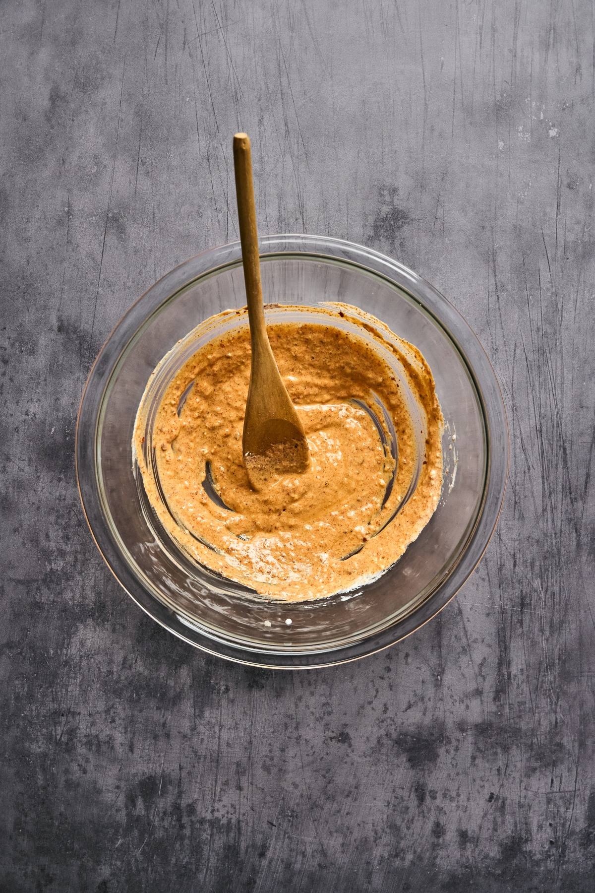 A glass bowl with brown batter and a wooden spoon on a gray textured surface.