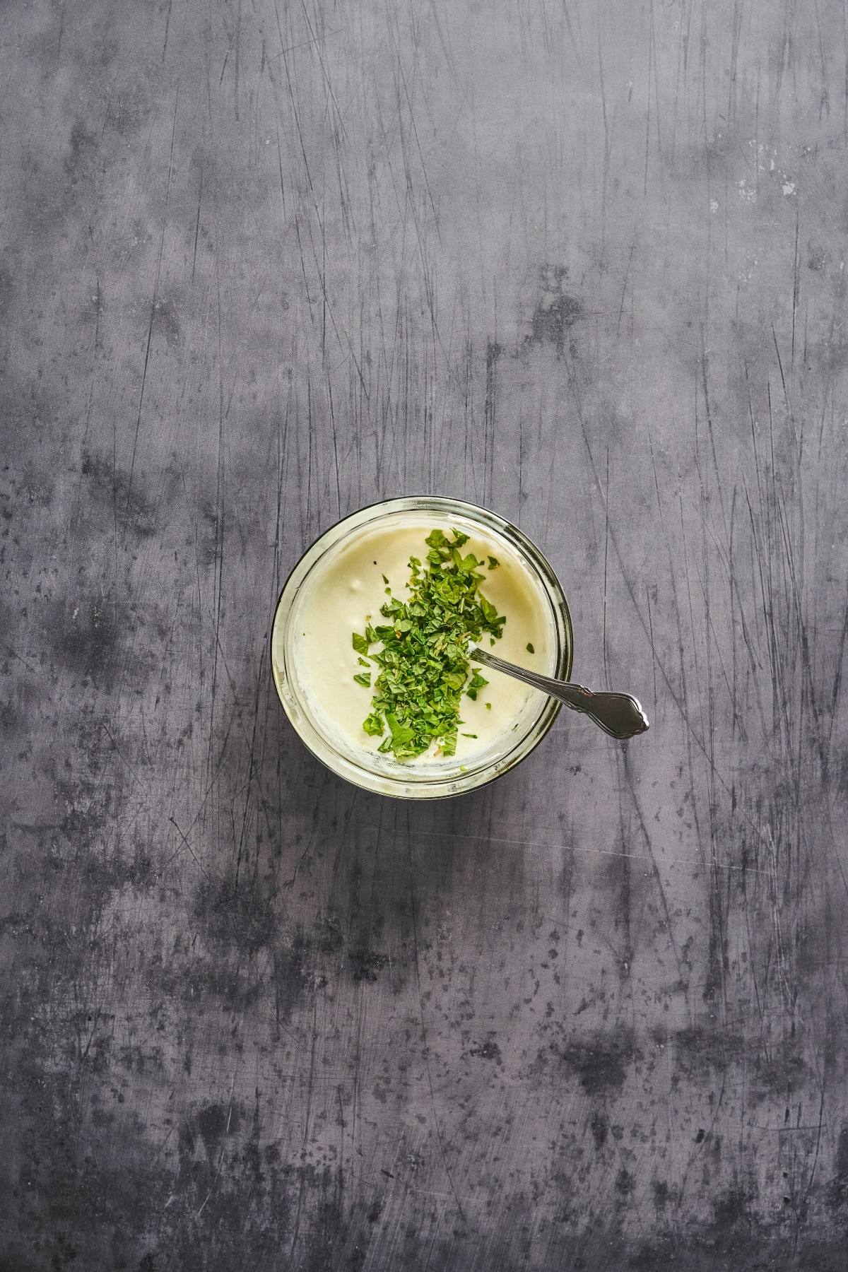 A bowl of creamy dip topped with chopped herbs and a spoon, on a textured gray surface.