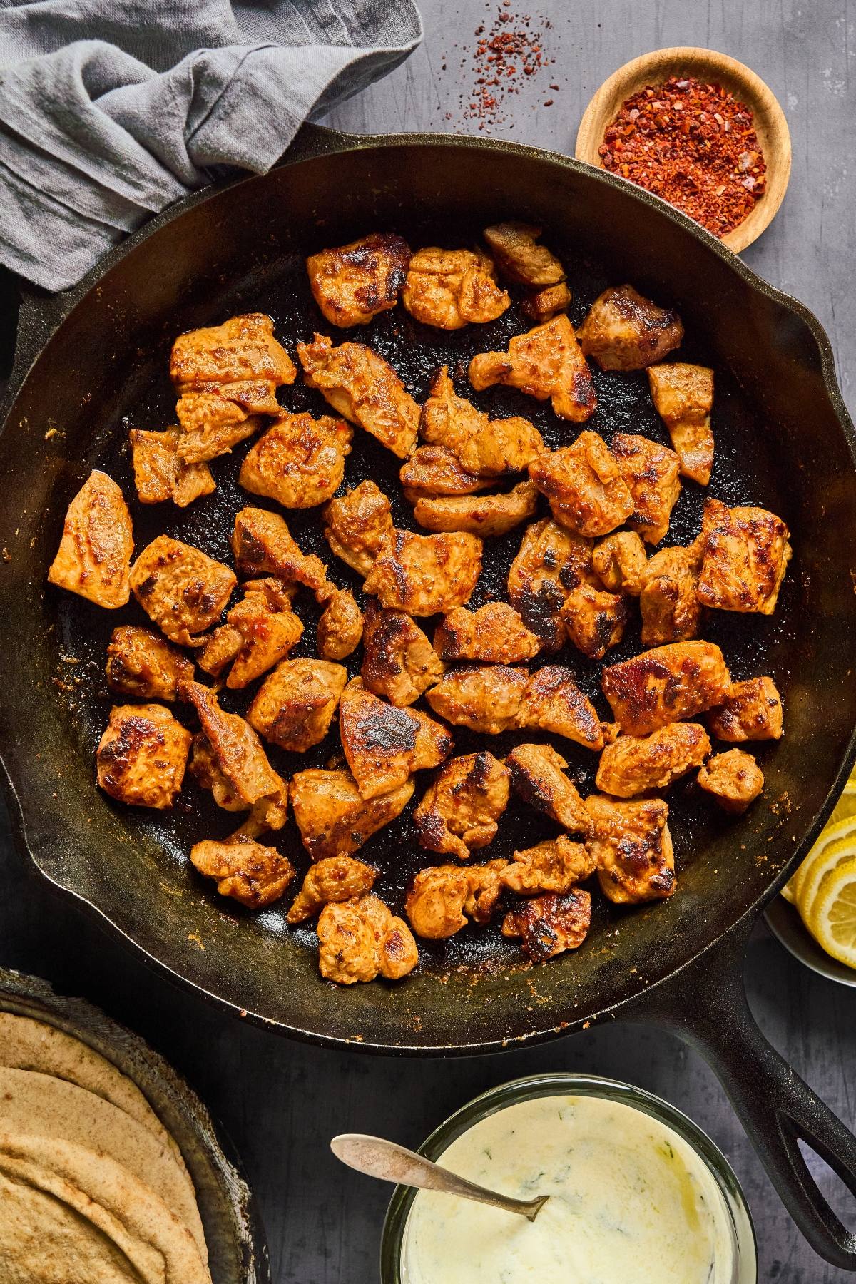 Chunks of spiced chicken cooking in a cast iron skillet, with sauce, pita, and seasonings nearby.