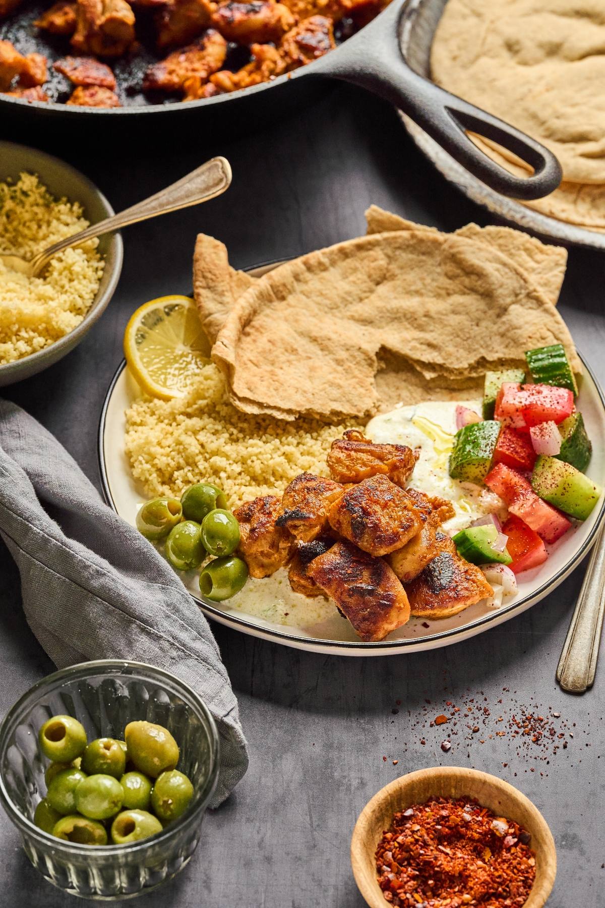 Plate with grilled chicken, pita bread, couscous, olives, tzatziki, and cucumber-tomato salad.
