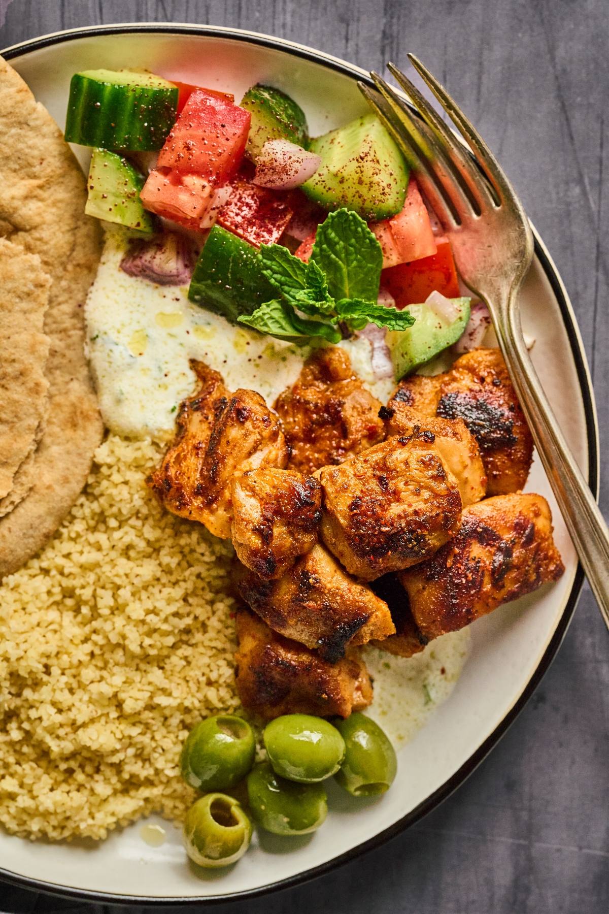 A plate with grilled chicken, couscous, pita bread, olives, salad, and yogurt sauce, with a fork on the side.