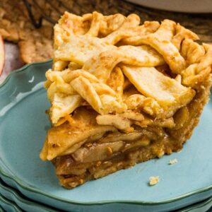 A slice of apple cheddar pie with a golden lattice crust sits on stacked blue plates next to sliced apples and forks.