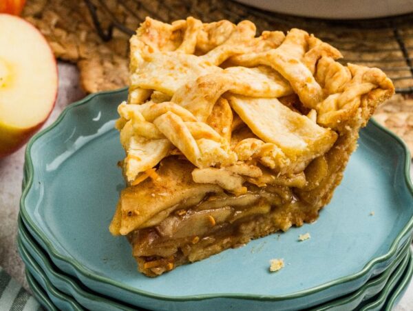A slice of apple cheddar pie with a golden lattice crust sits on stacked blue plates next to sliced apples and forks.