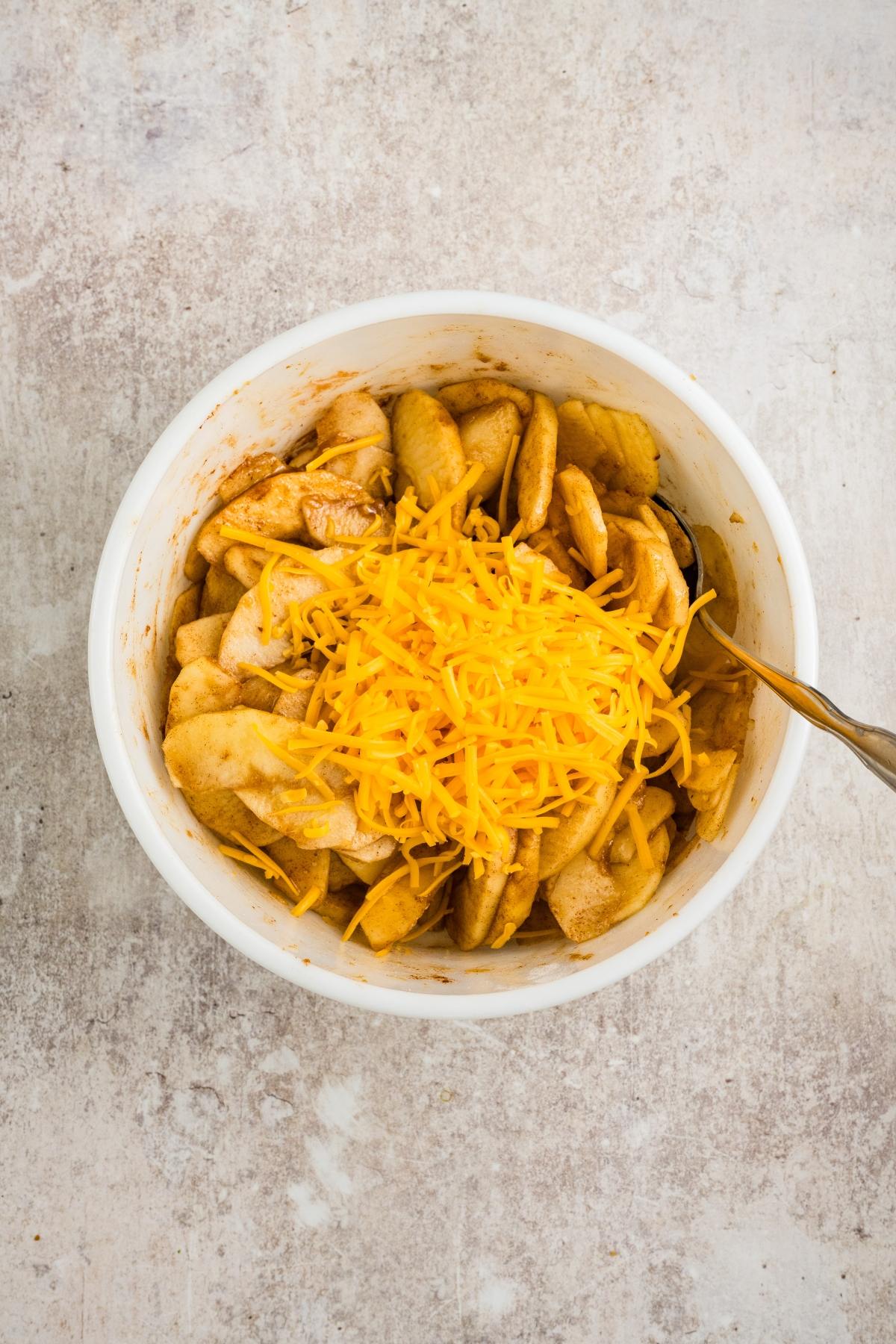 Sliced apples with shredded cheddar cheese and spices in a white bowl on a light surface.
