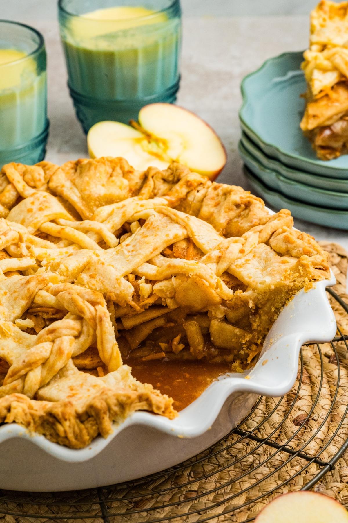 Sliced apple pie with lattice crust in a white dish, with apple slices and drinks in the background.