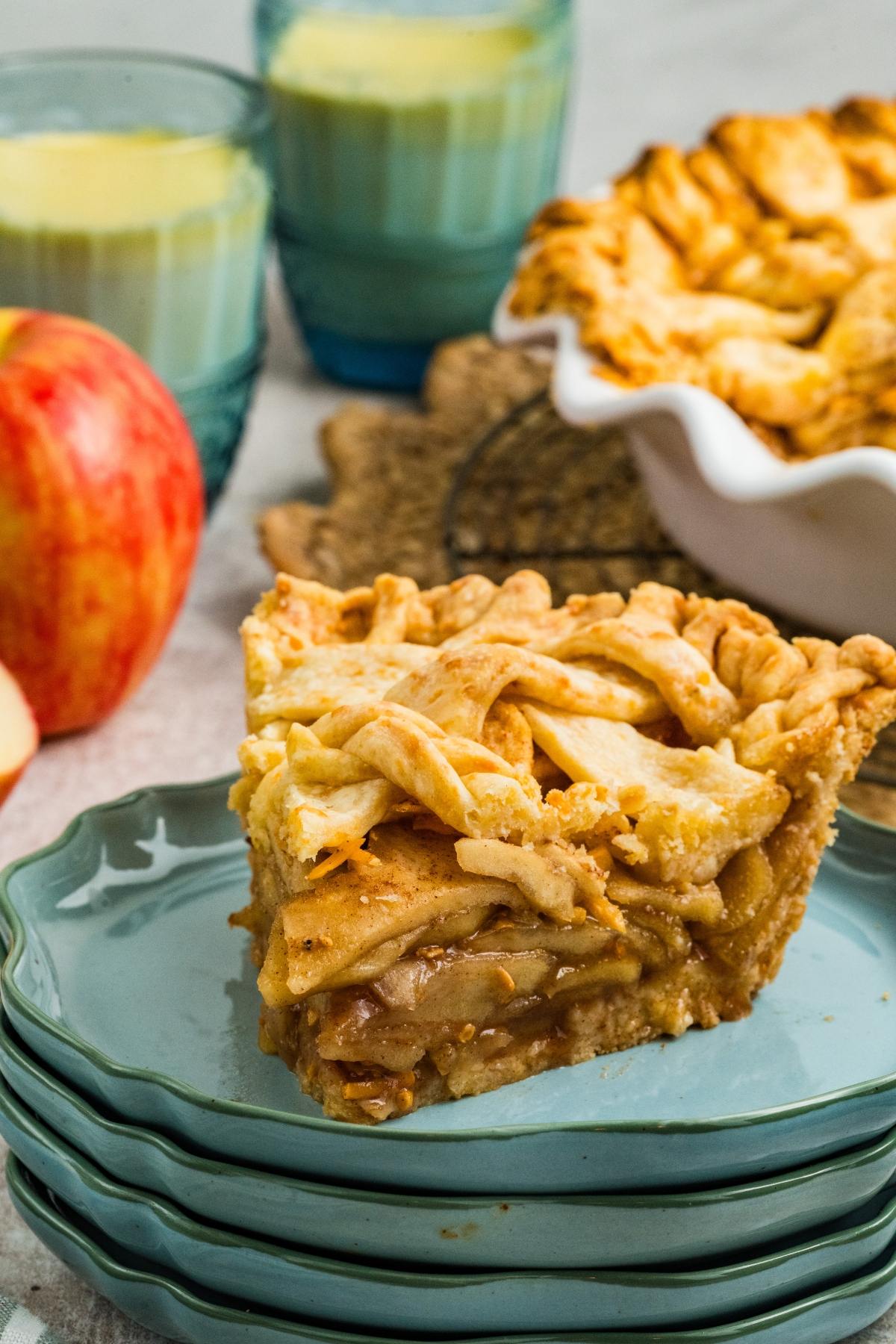 A slice of apple pie on stacked plates with a whole pie, apples, and drinks in the background.