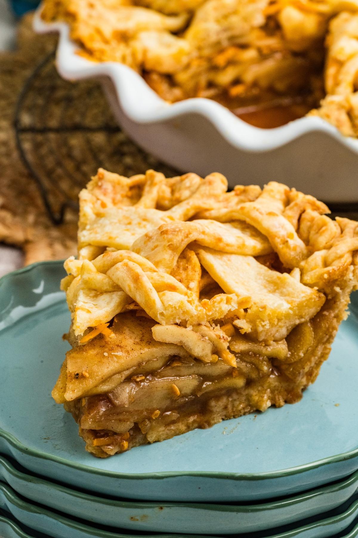 A slice of lattice-topped apple pie on a stack of blue plates, with the rest of the pie in the background.