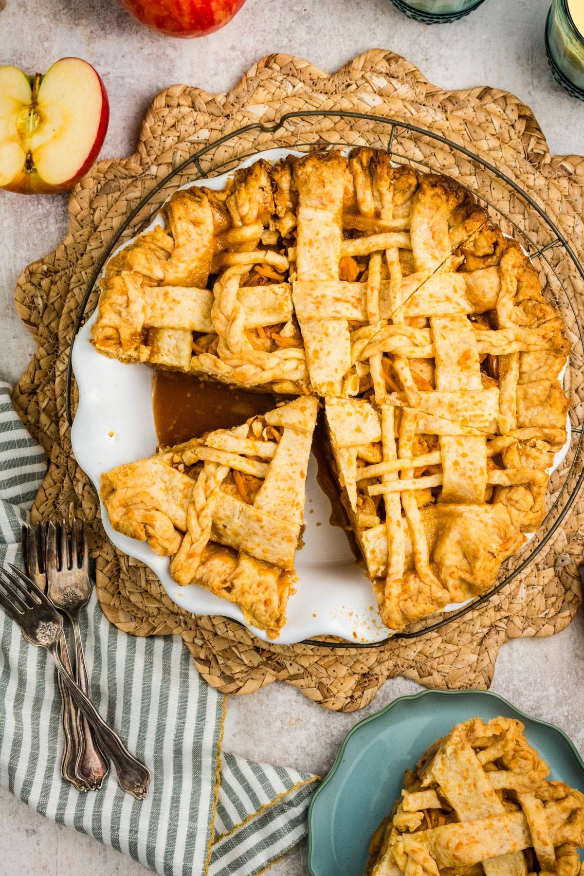 A lattice-topped apple pie with two slices cut out, on a white plate, next to apples and forks.
