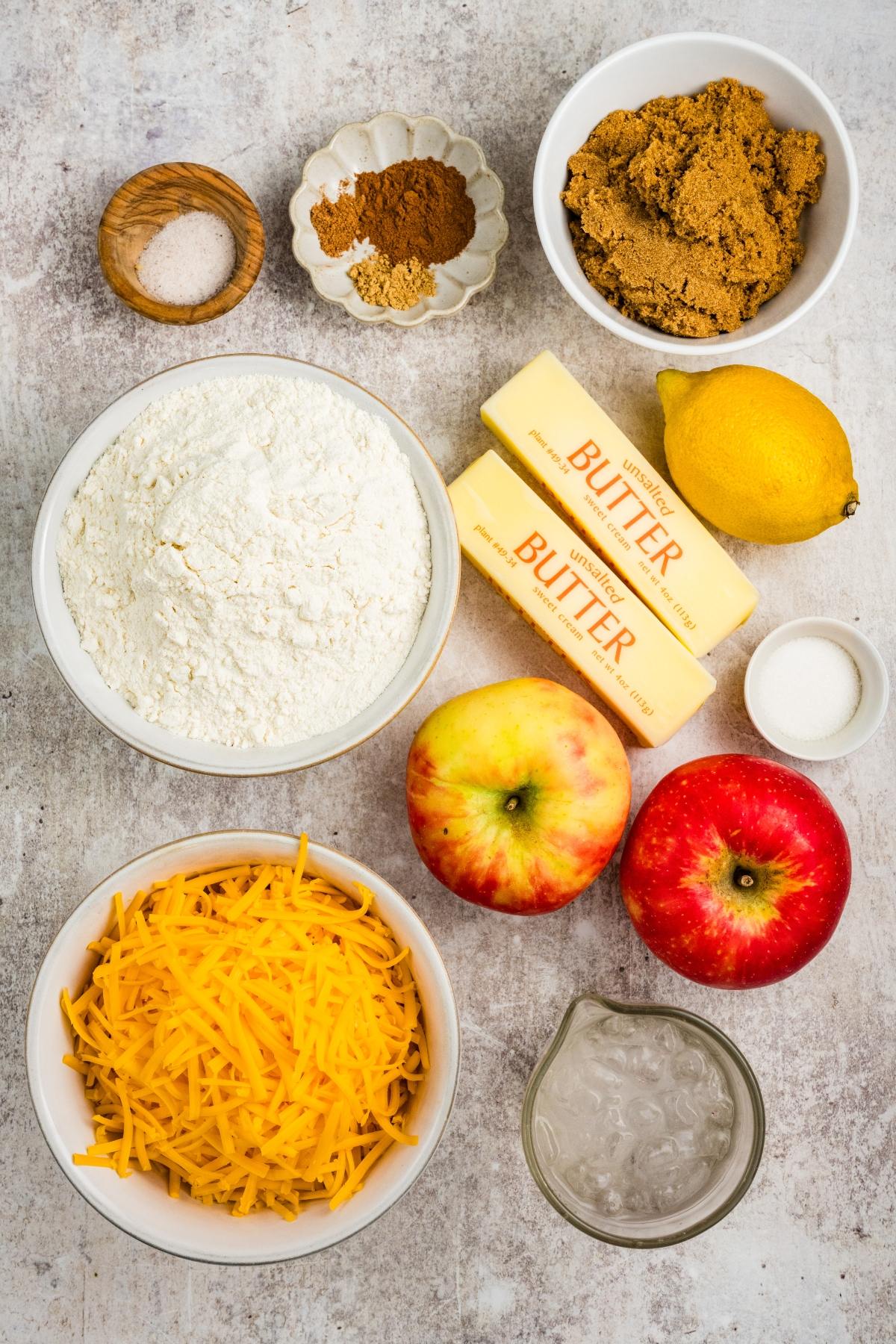 Ingredients for baking: flour, butter, cheese, apples, lemon, sugar, brown sugar, spices, and salt on a counter.