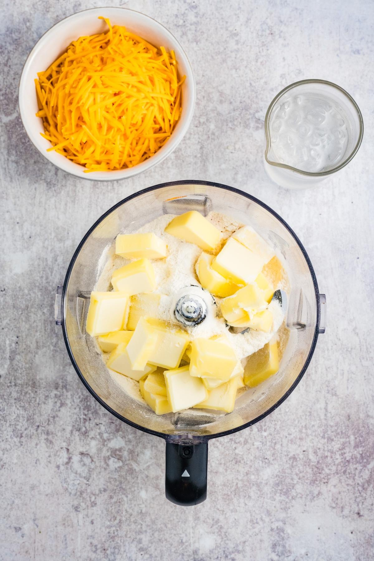 Food processor with flour and butter, a bowl of shredded cheese, and a cup of water on a countertop.