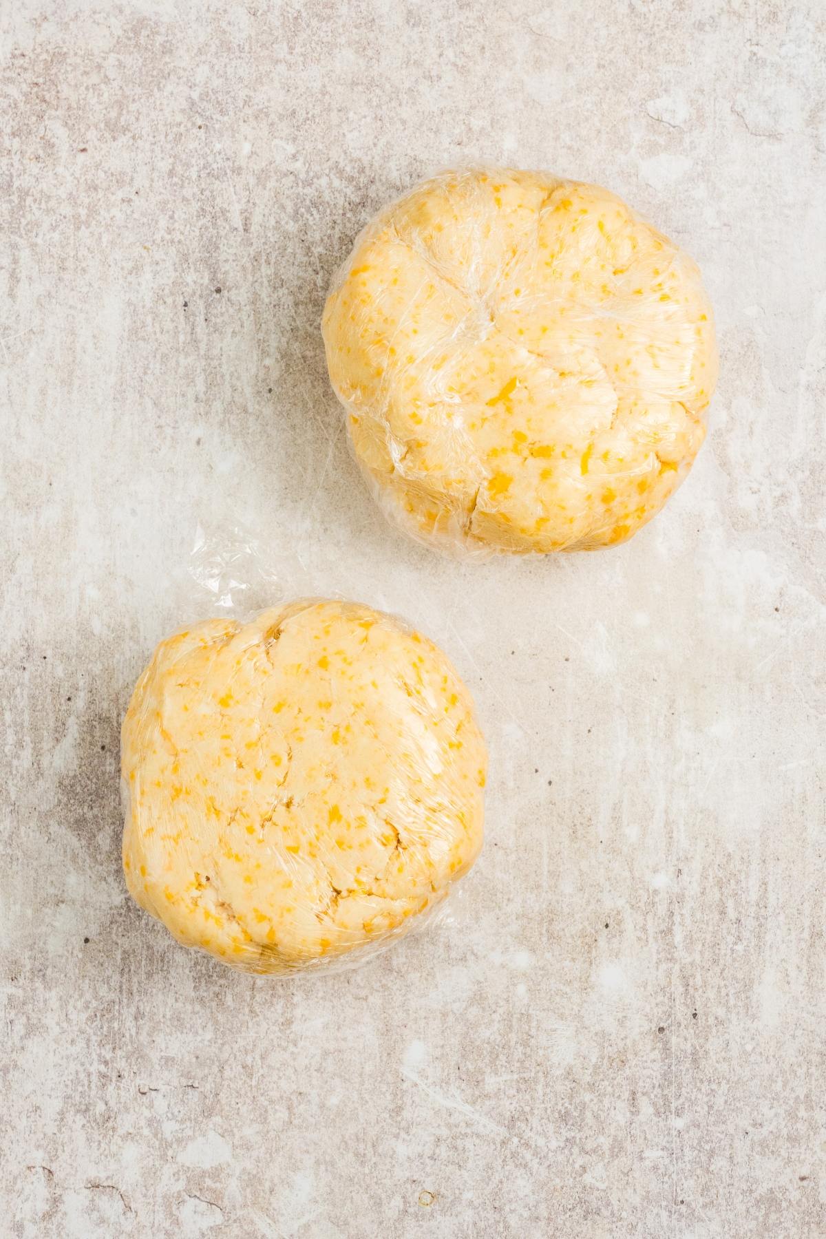 Two round discs of dough wrapped in plastic wrap on a light textured surface.