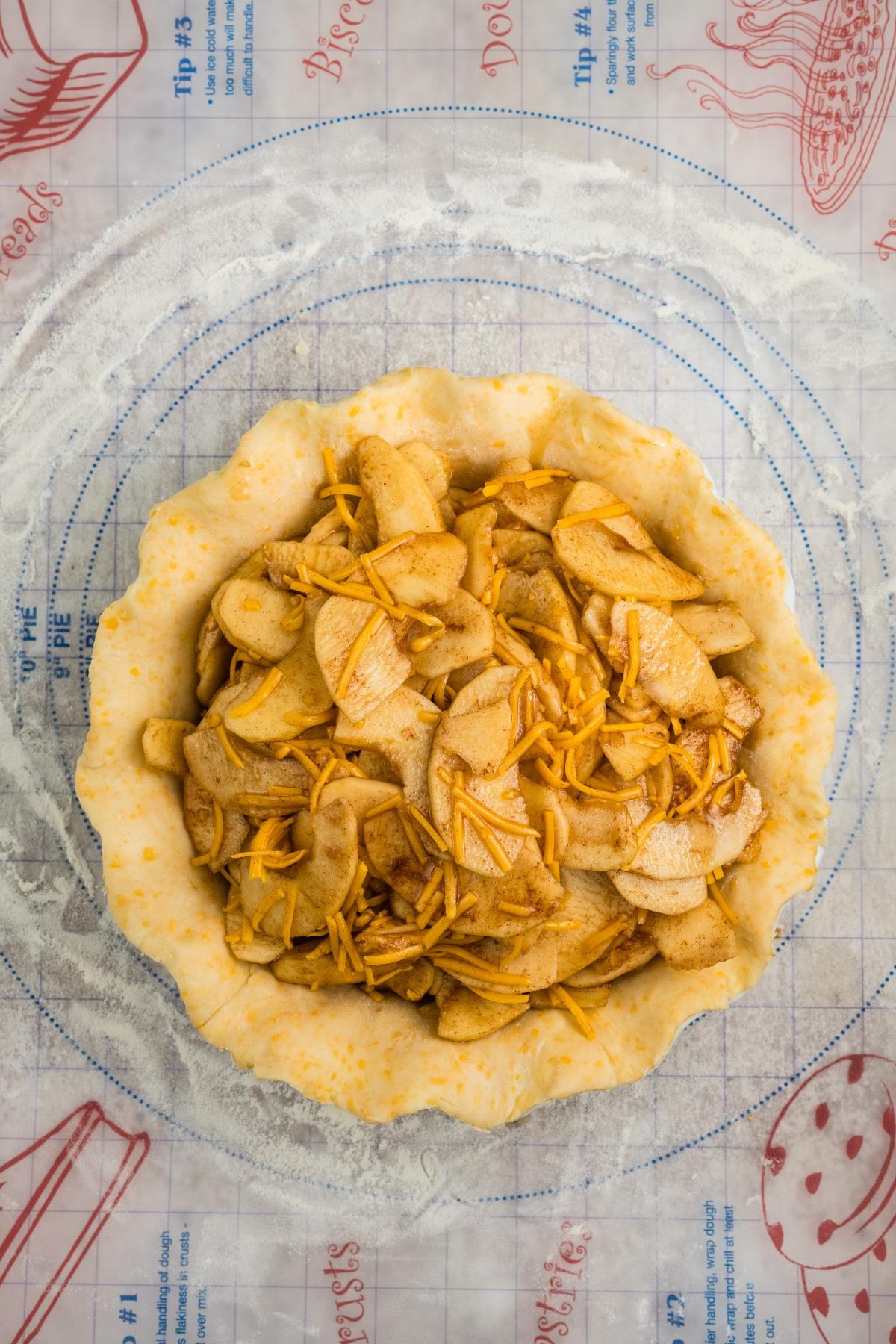Unbaked apple pie with sliced apples and shredded cheese in a pie crust on a pastry mat.