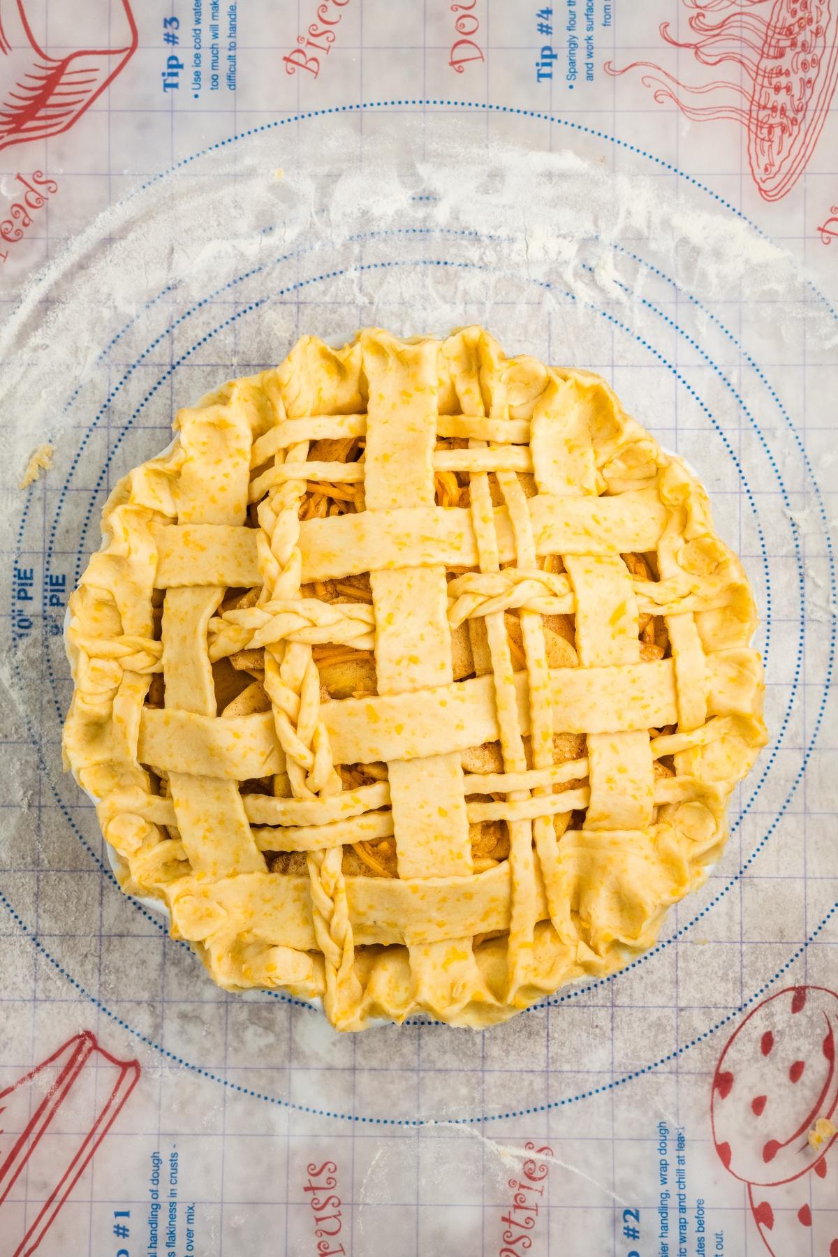 Unbaked lattice-top pie with braided crust on a floured pastry mat with baking guides and pie illustrations.