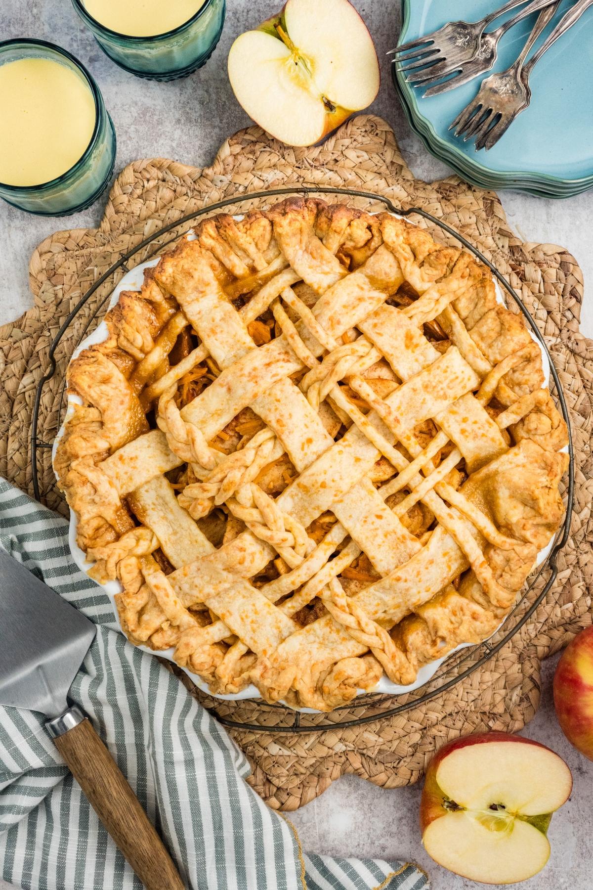 Lattice-topped apple pie on a woven mat, surrounded by apple slices, forks, and glasses of milk.