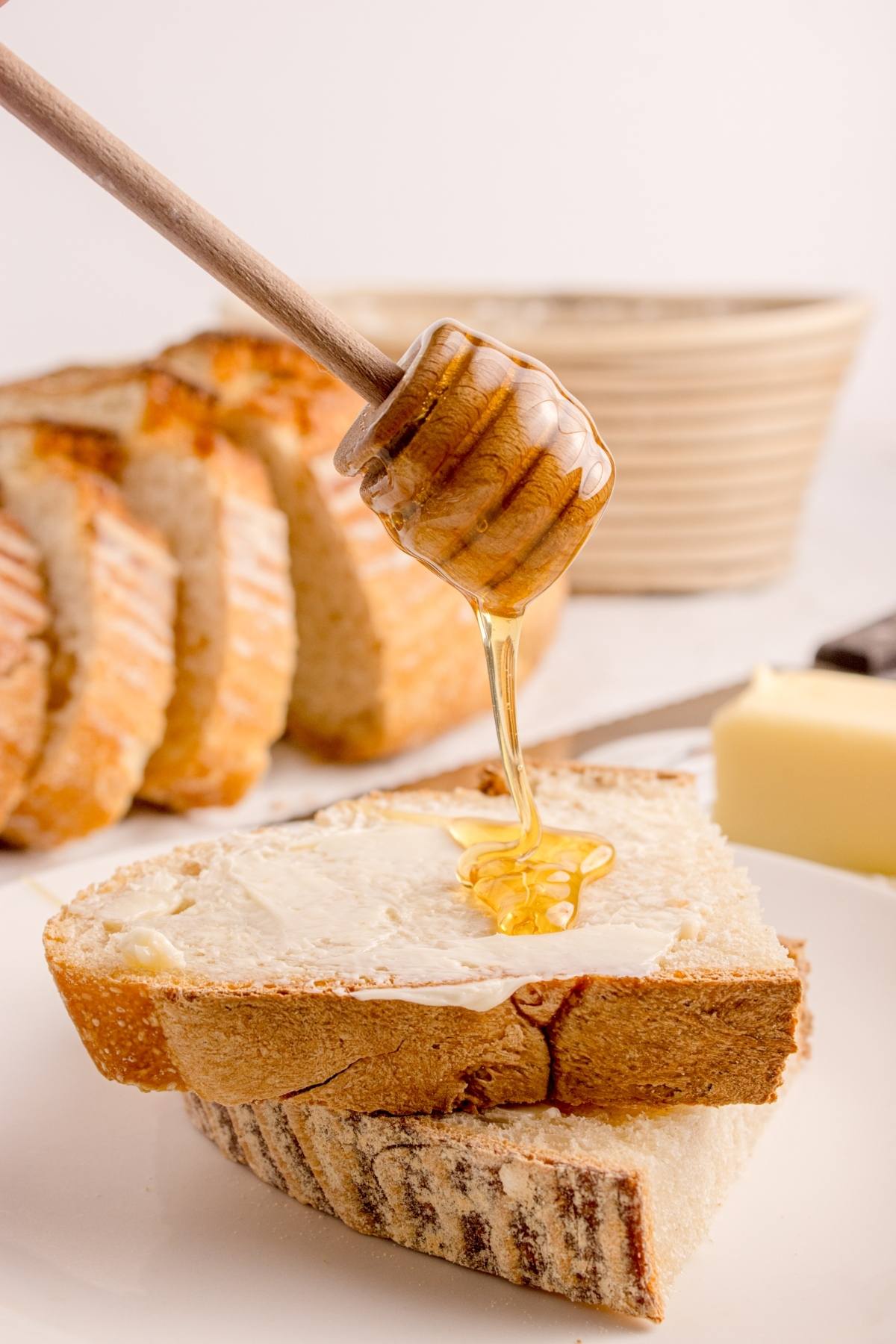 A honey dipper drizzles honey onto buttered bread slices, with more bread and butter in the background.