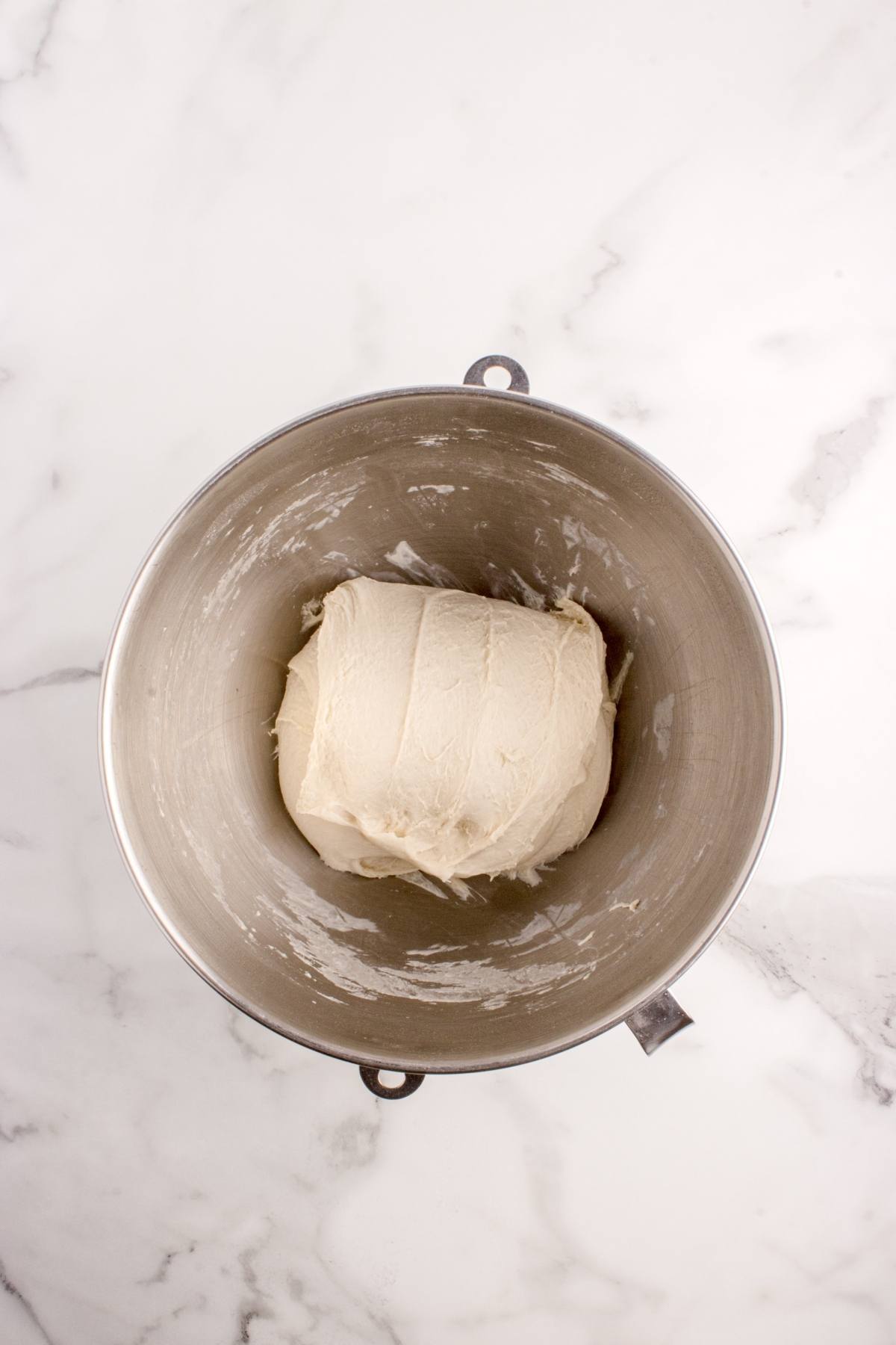 A ball of dough in a metal mixing bowl on a white marble surface.