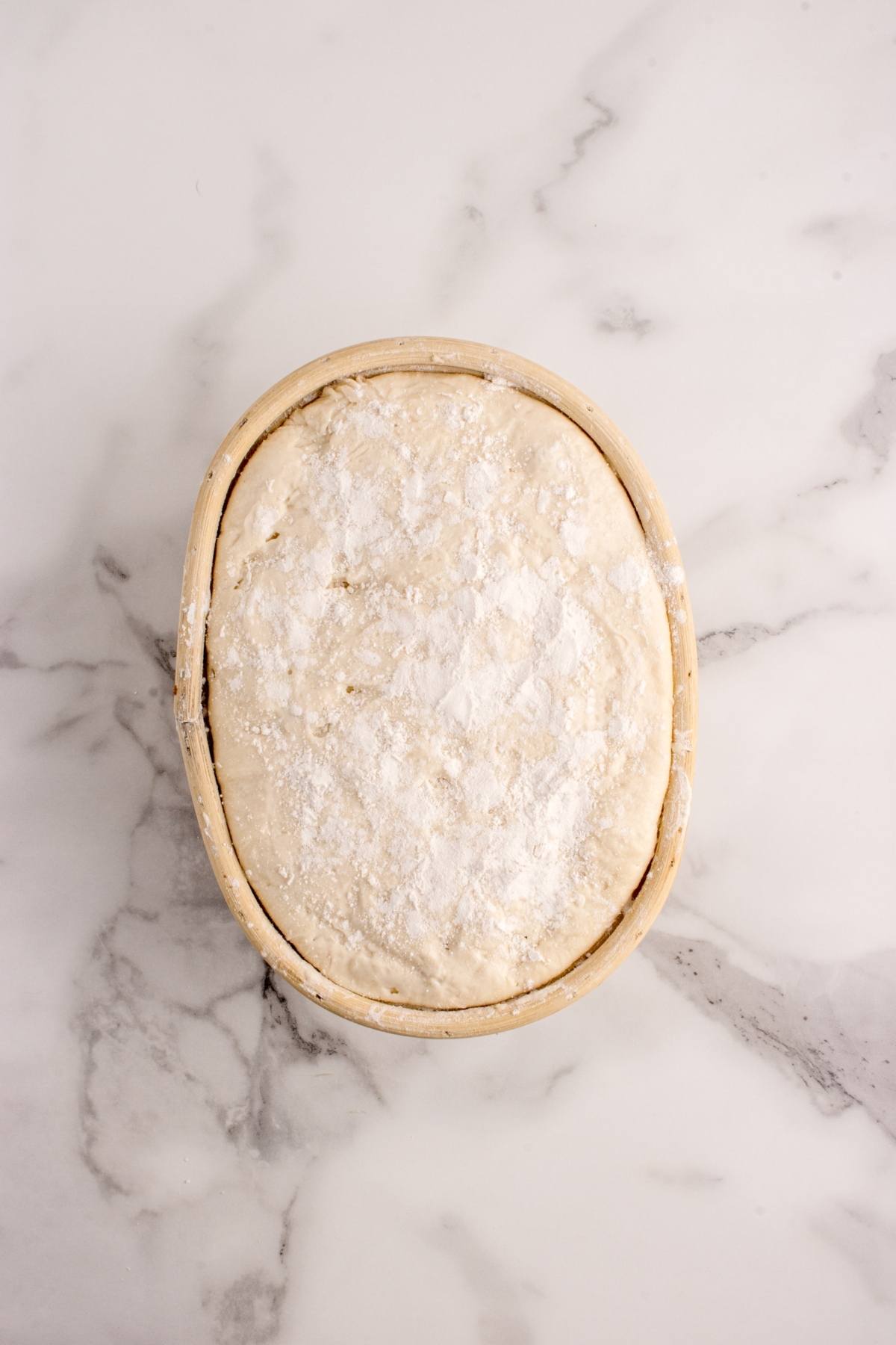 Oval basket with floured bread dough rising on a white marble surface.