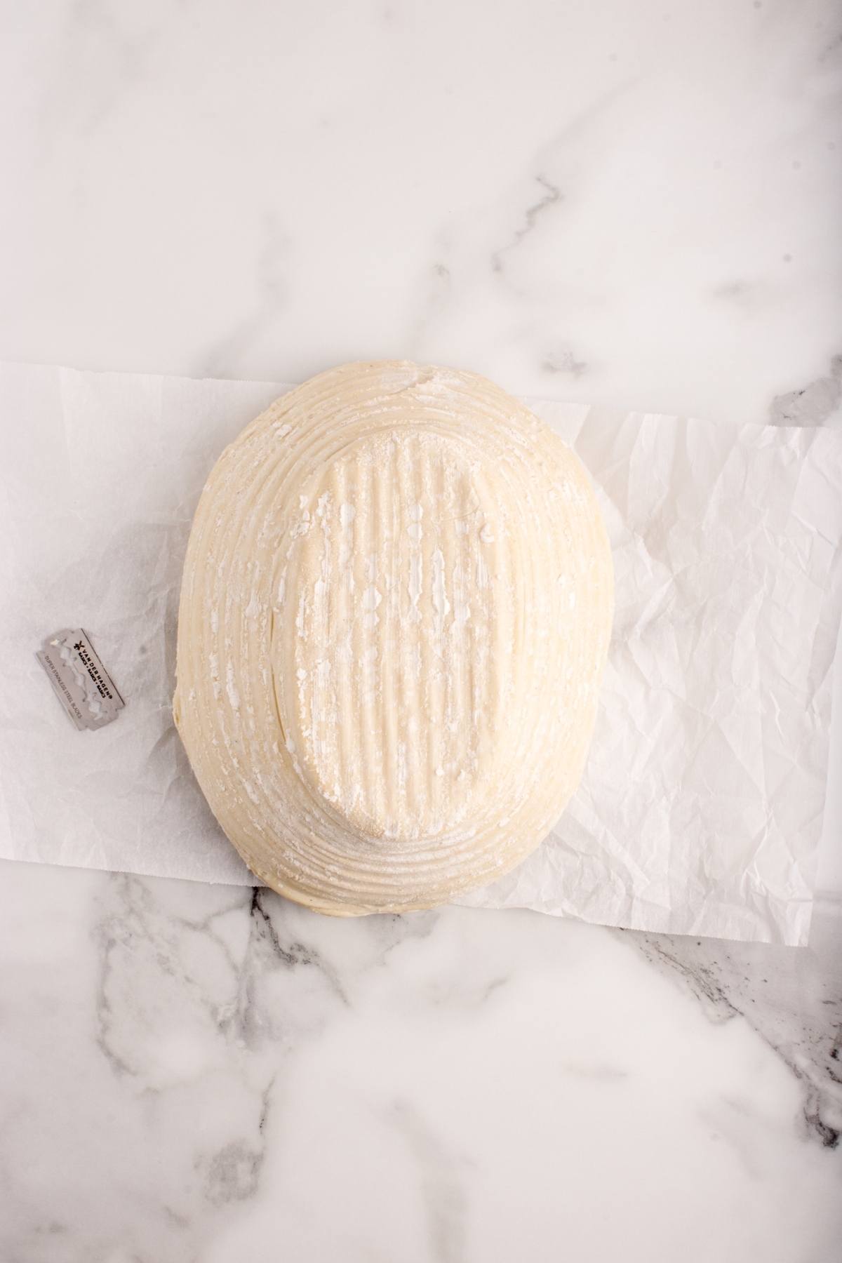 A round, floured bread dough on parchment paper, resting on a white marble surface.