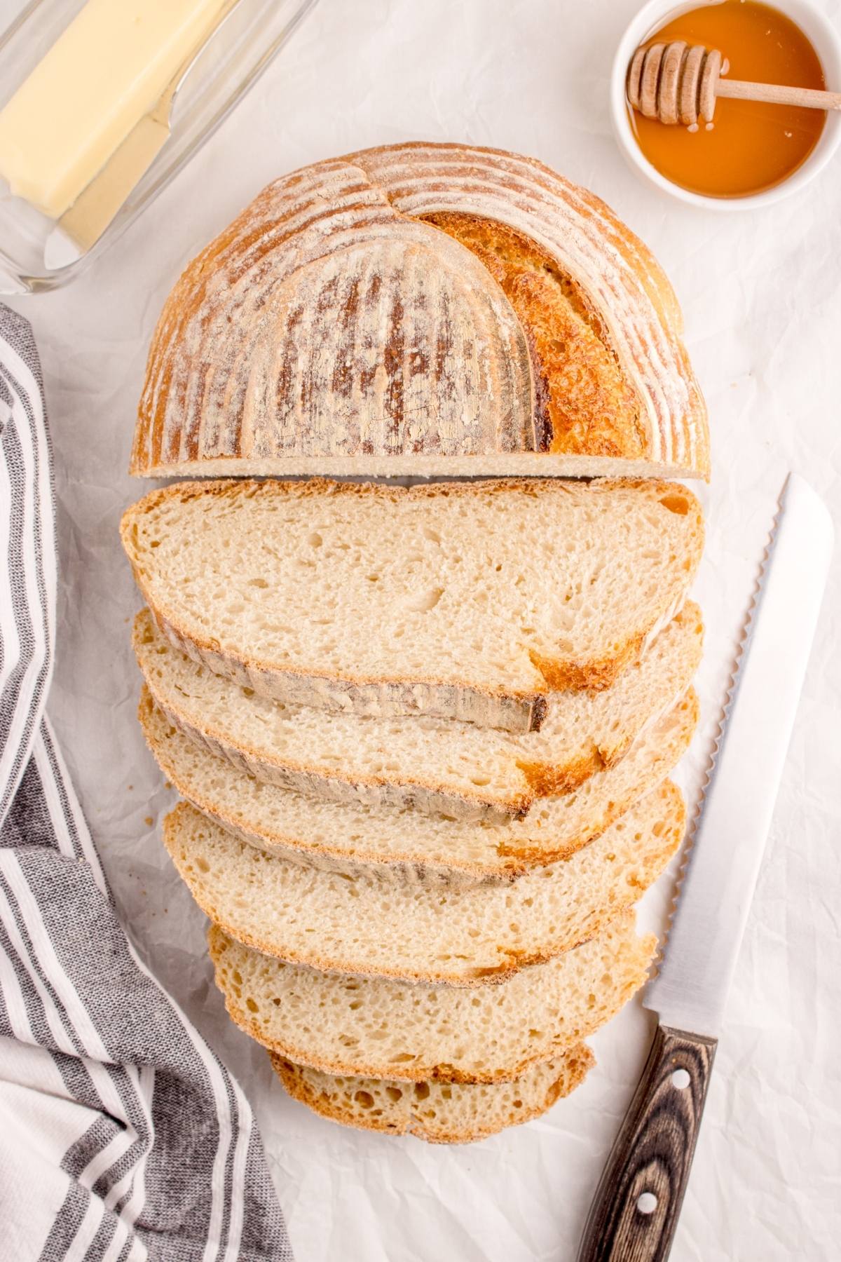 Sliced round loaf of bread with a knife, honey, and butter on a white surface.