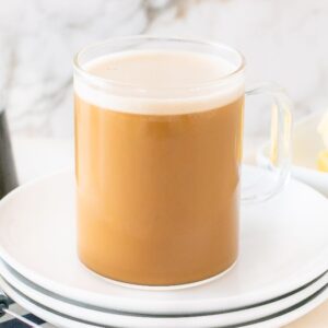 A glass mug of creamy bulletproof coffee sits on stacked plates, with syrup and butter in the background.