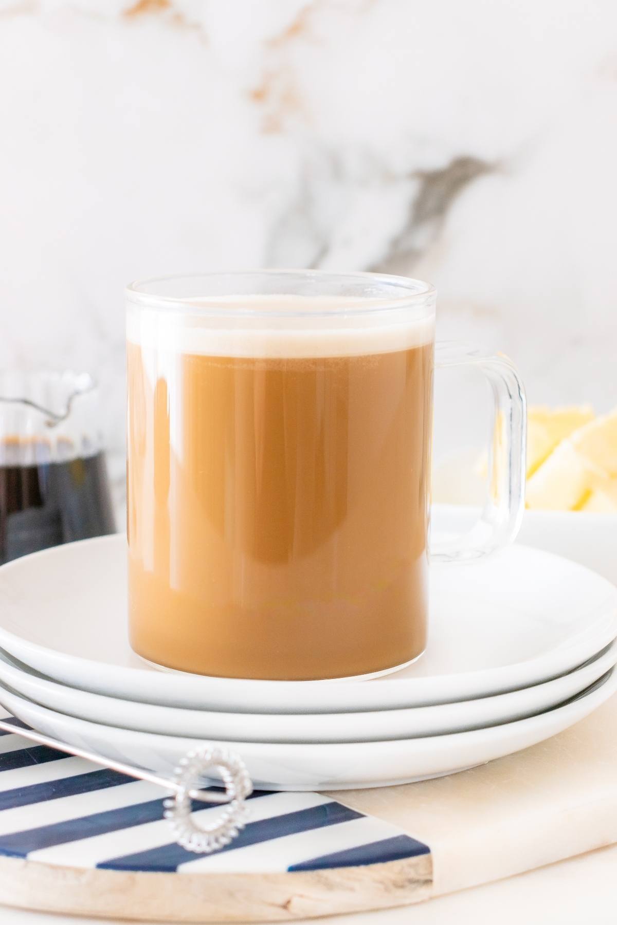 A glass mug of coffee with milk on stacked white plates, next to a metal whisk and a striped napkin.