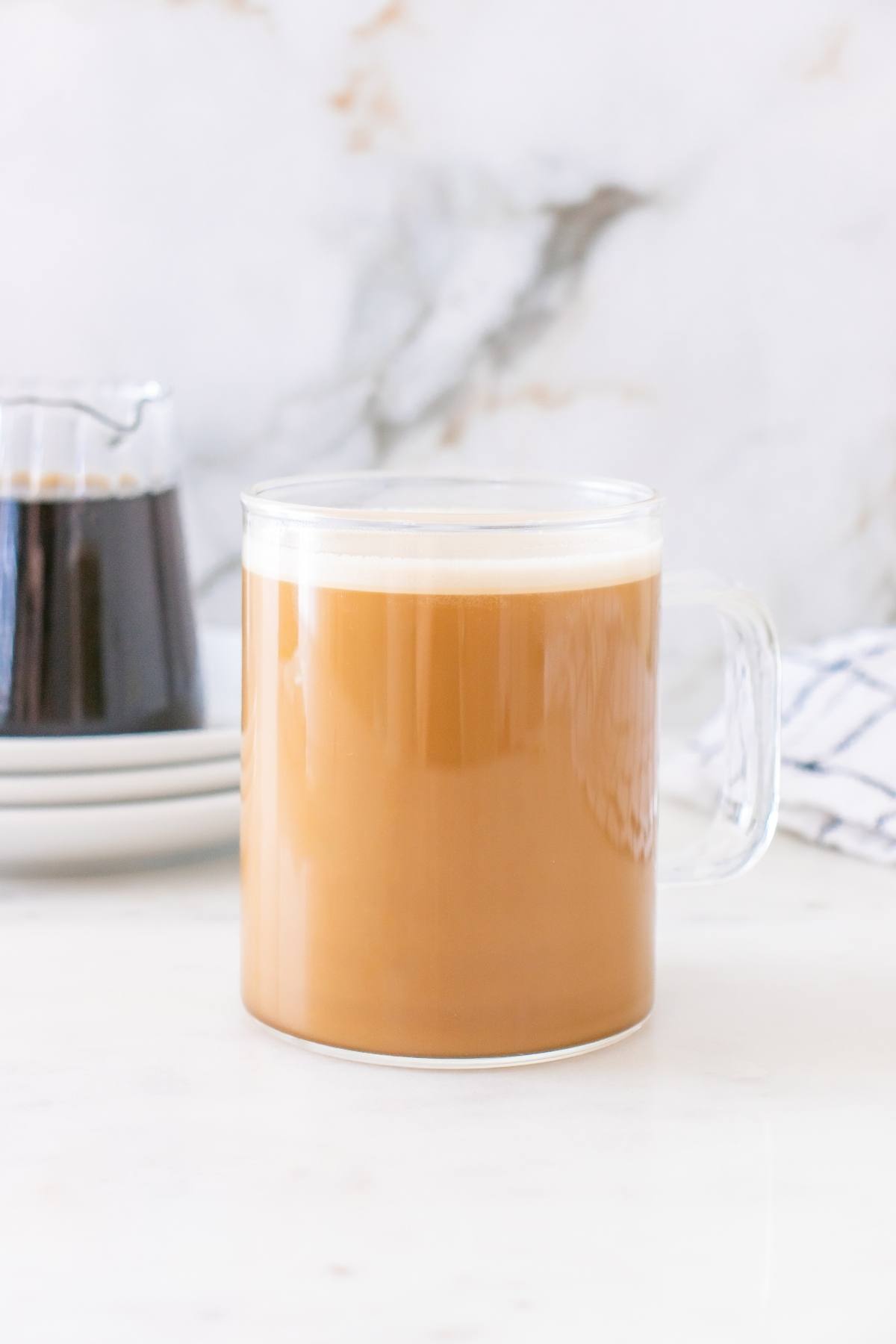Clear glass mug filled with creamy coffee on a white surface, with coffee pot and towel in background.