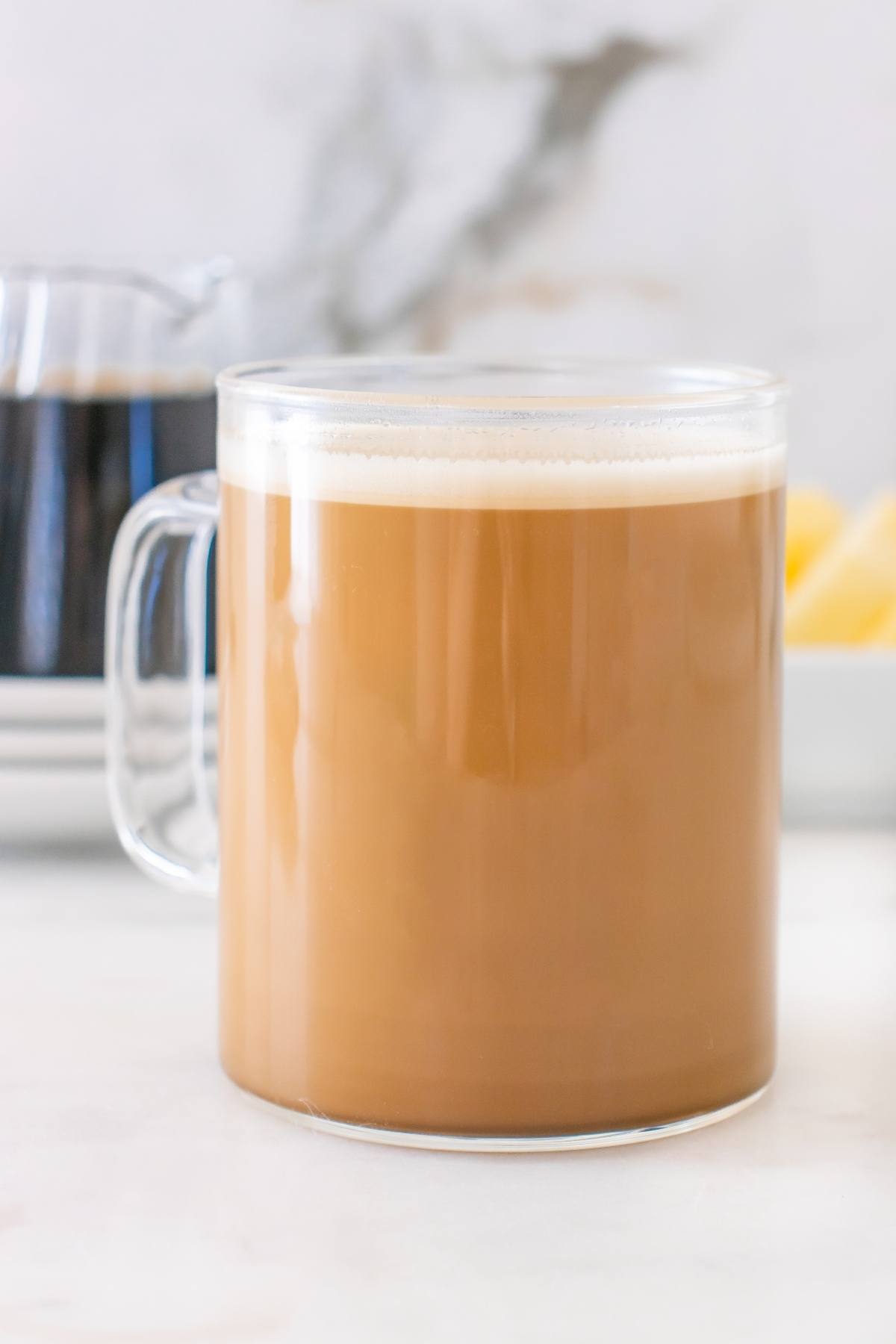 A clear glass mug filled with creamy coffee sits on a white surface with a blurred background.