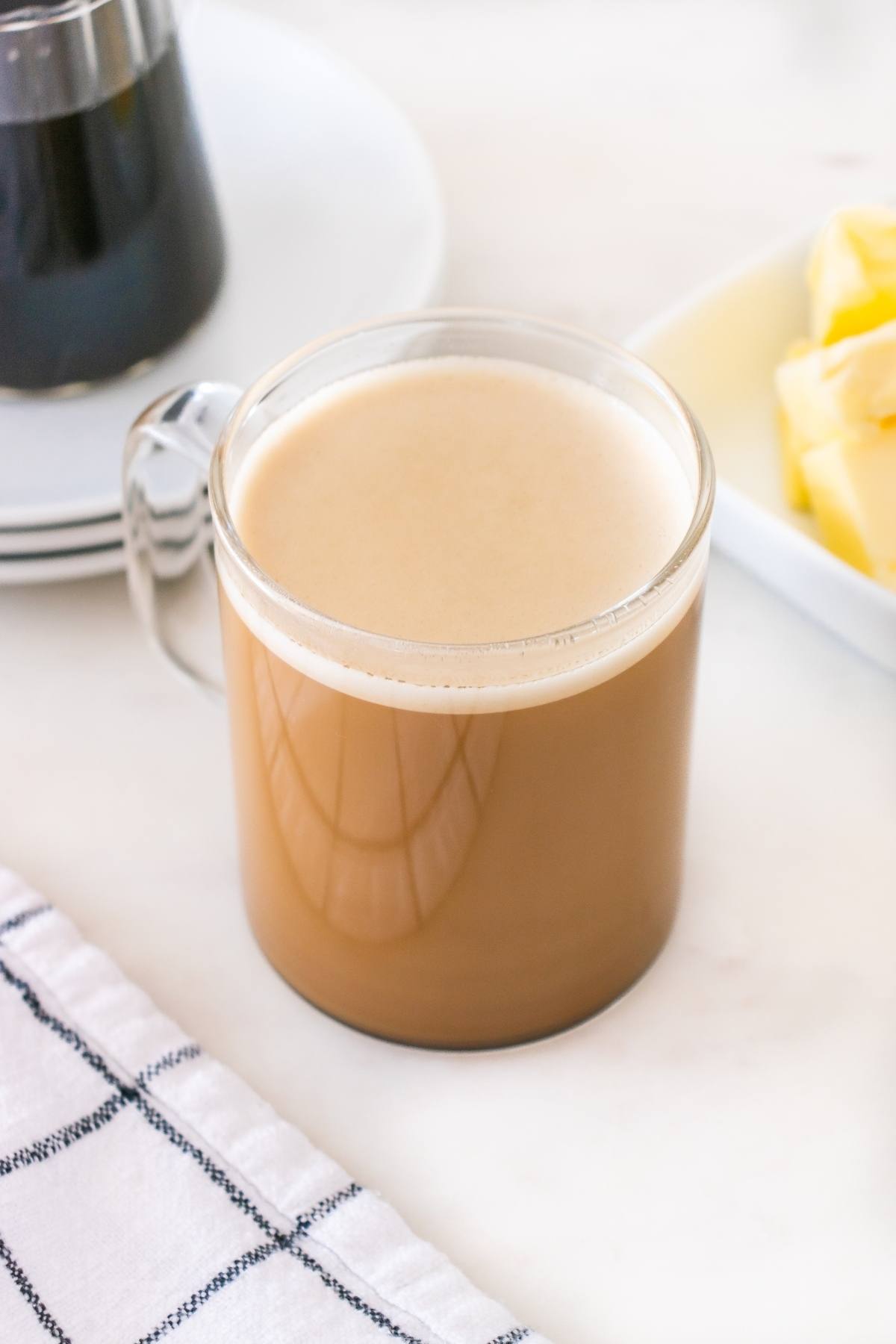 A glass mug of creamy coffee sits on a white table next to a plate of butter and a checkered napkin.