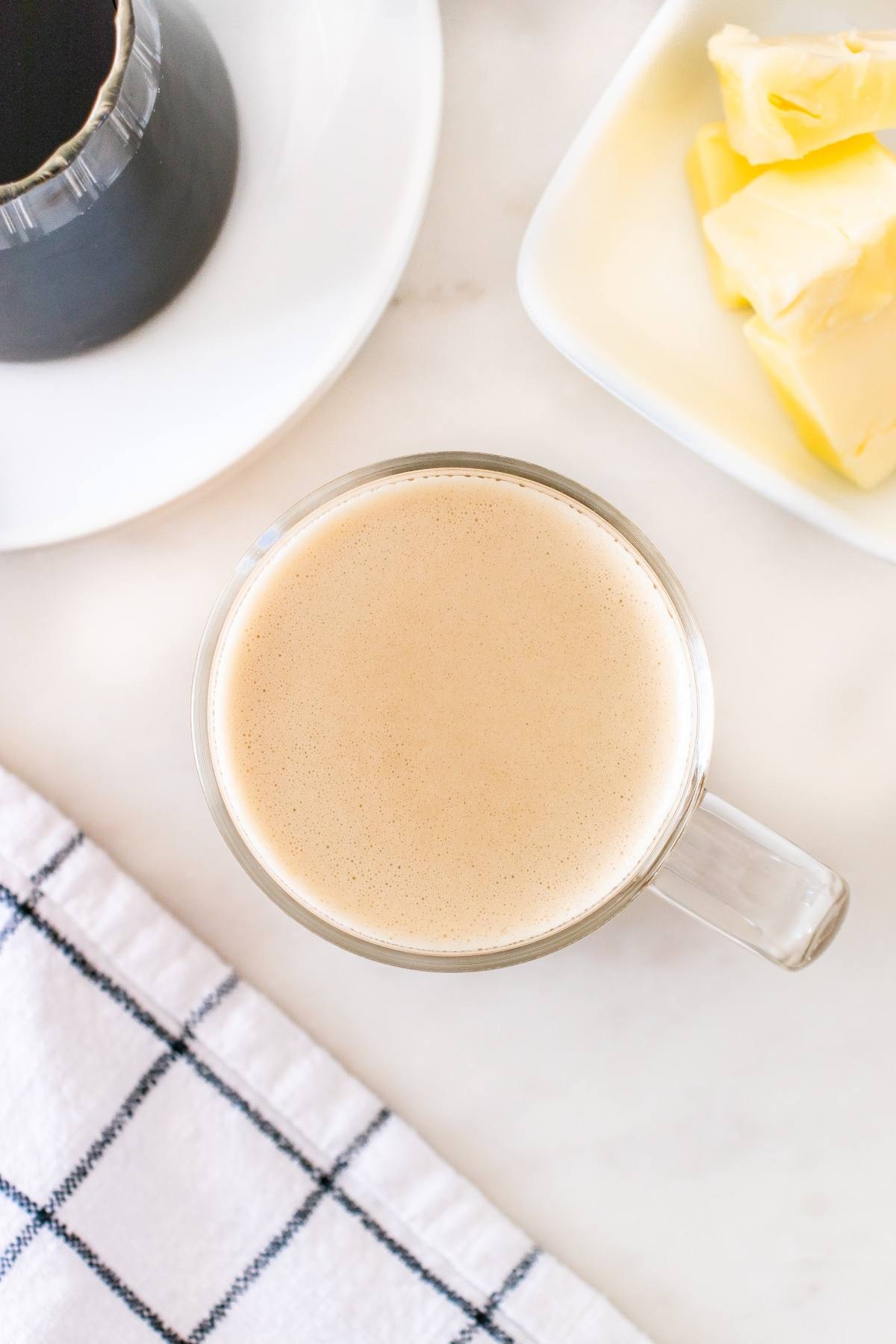 A glass mug of creamy coffee next to butter on a plate and a checkered towel on a white surface.