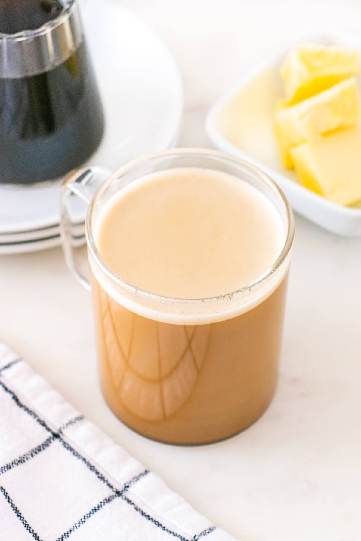 A glass mug of creamy coffee sits on a table near a dish of butter and a black and white checkered towel.