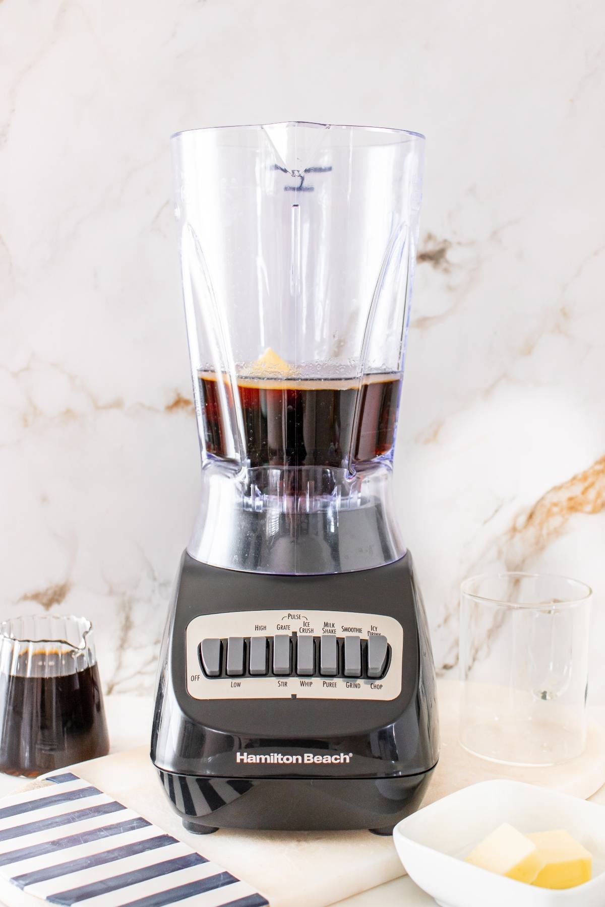 A blender with dark liquid and ice inside, on a counter beside a glass, a small pitcher, and a plate with ice cubes.