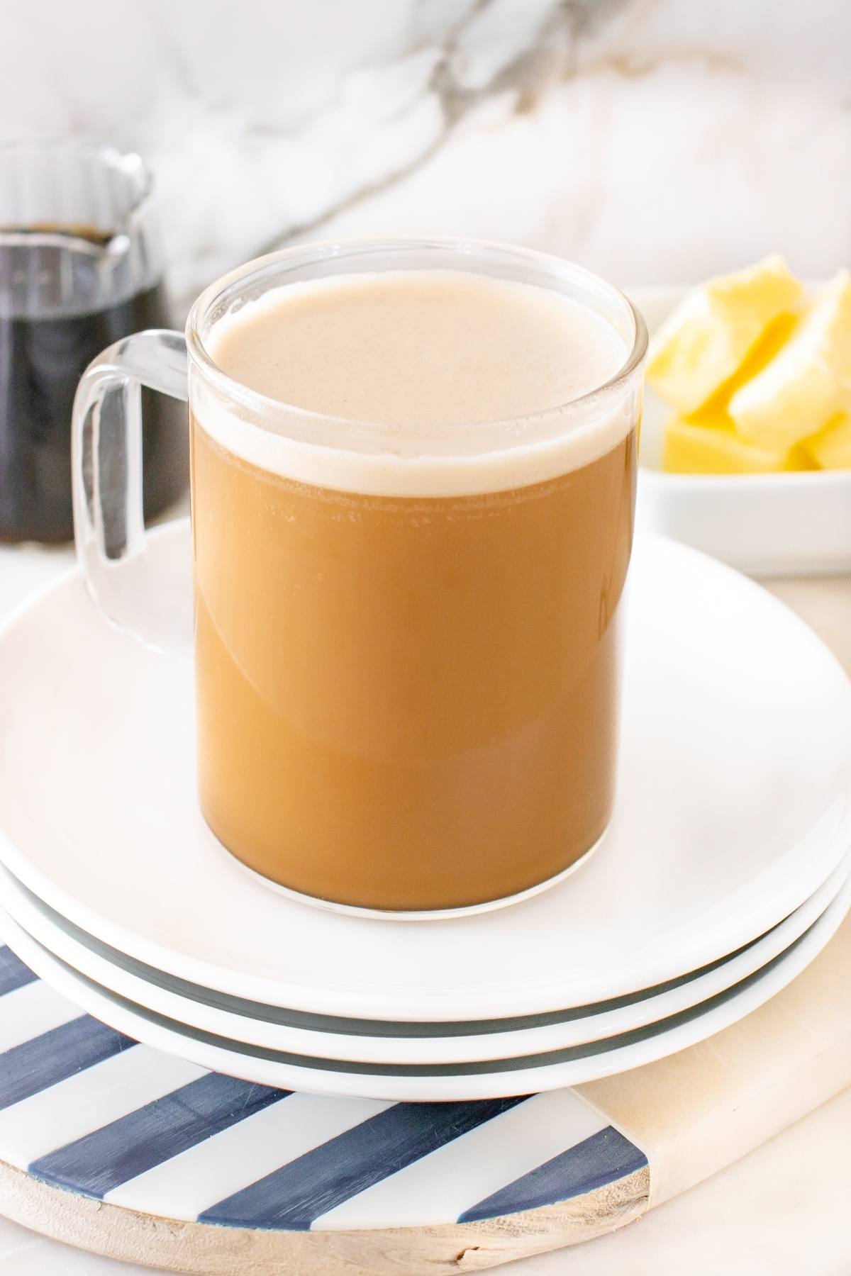Glass mug of creamy coffee on stacked white plates, with butter and a small jar of dark liquid in the background.