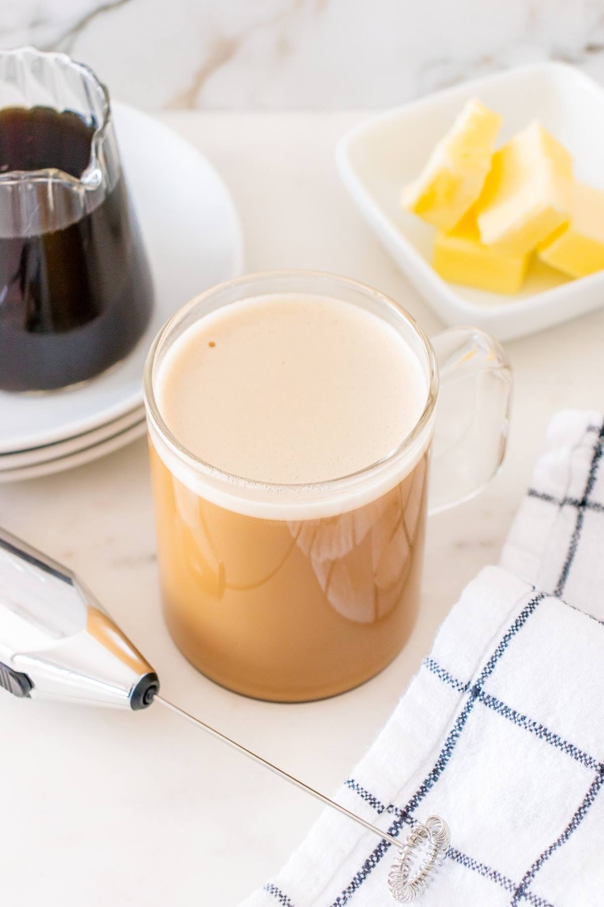 A glass mug with brown liquid and a spoon next to butter and a plate of butter.