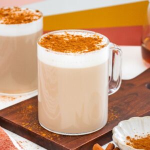 Two frothy chai lattes with cinnamon on top, one on a wooden board, next to honey and cinnamon sticks—perfect for fans of café con miel.