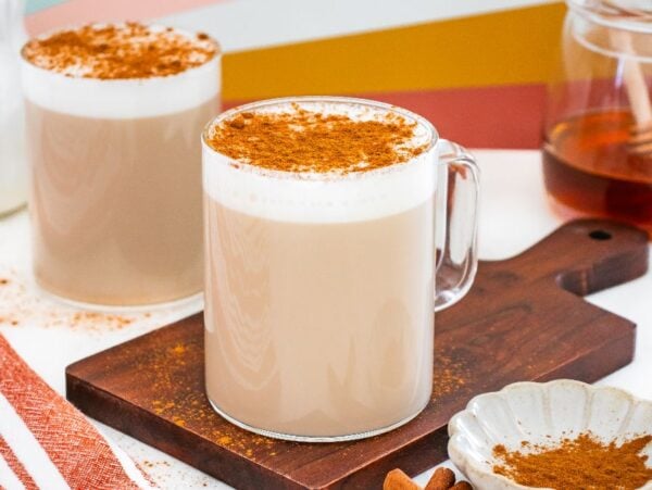 Two frothy chai lattes with cinnamon on top, one on a wooden board, next to honey and cinnamon sticks—perfect for fans of café con miel.