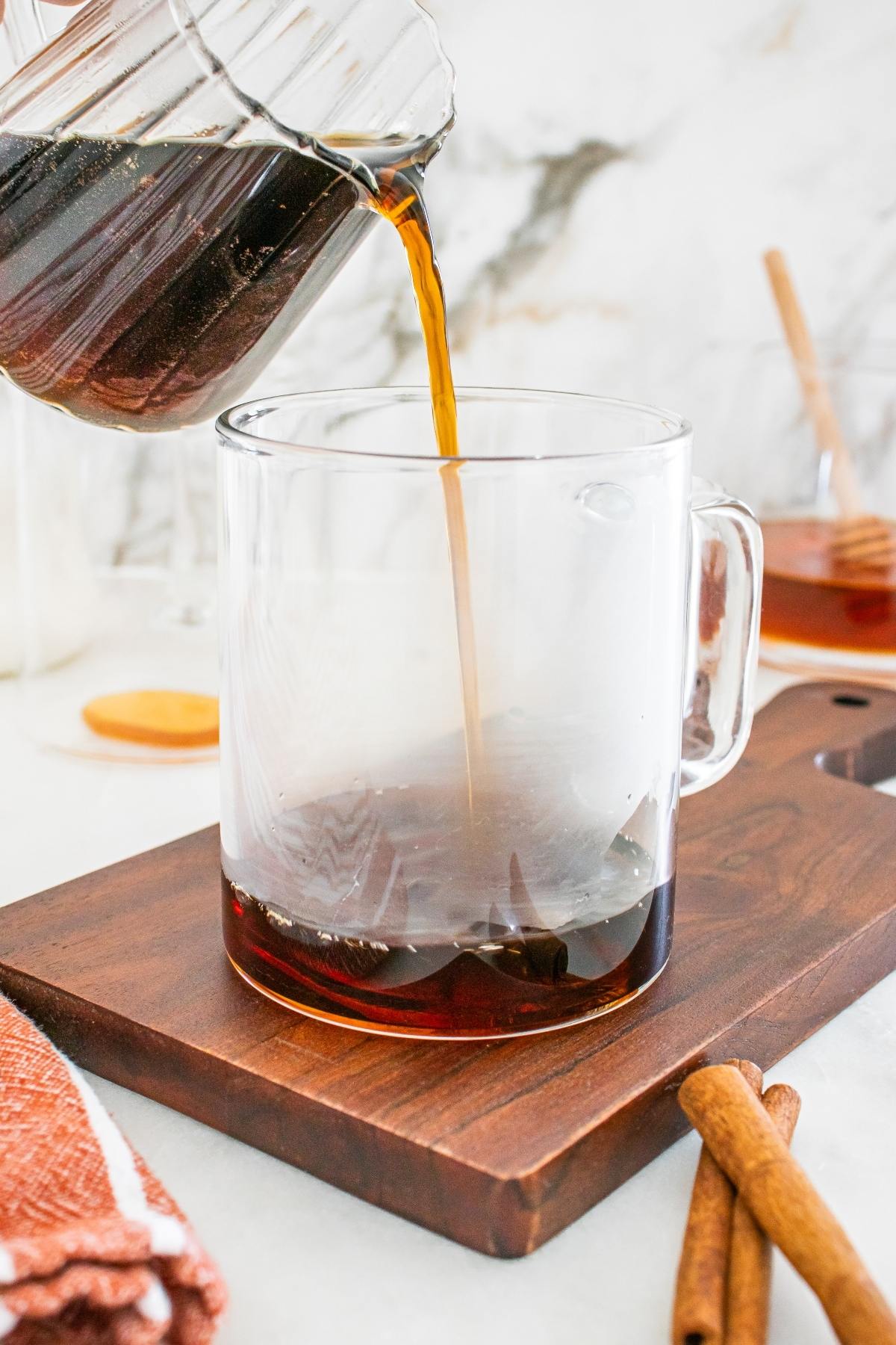Coffee being poured from a glass carafe into a clear mug on a wooden board with cinnamon sticks nearby.