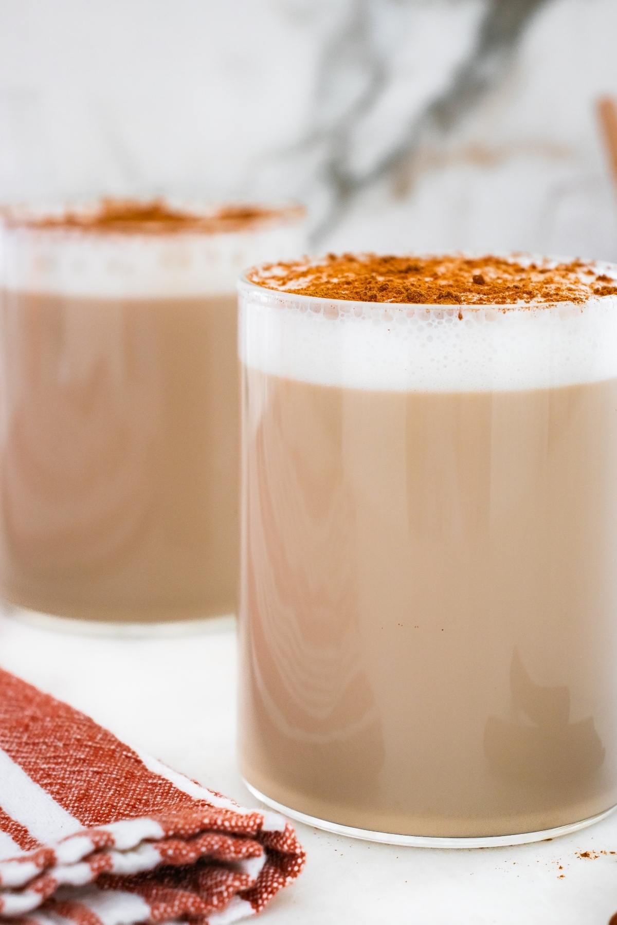 Two glasses of frothy chai latte topped with cinnamon, next to a red and white striped cloth.